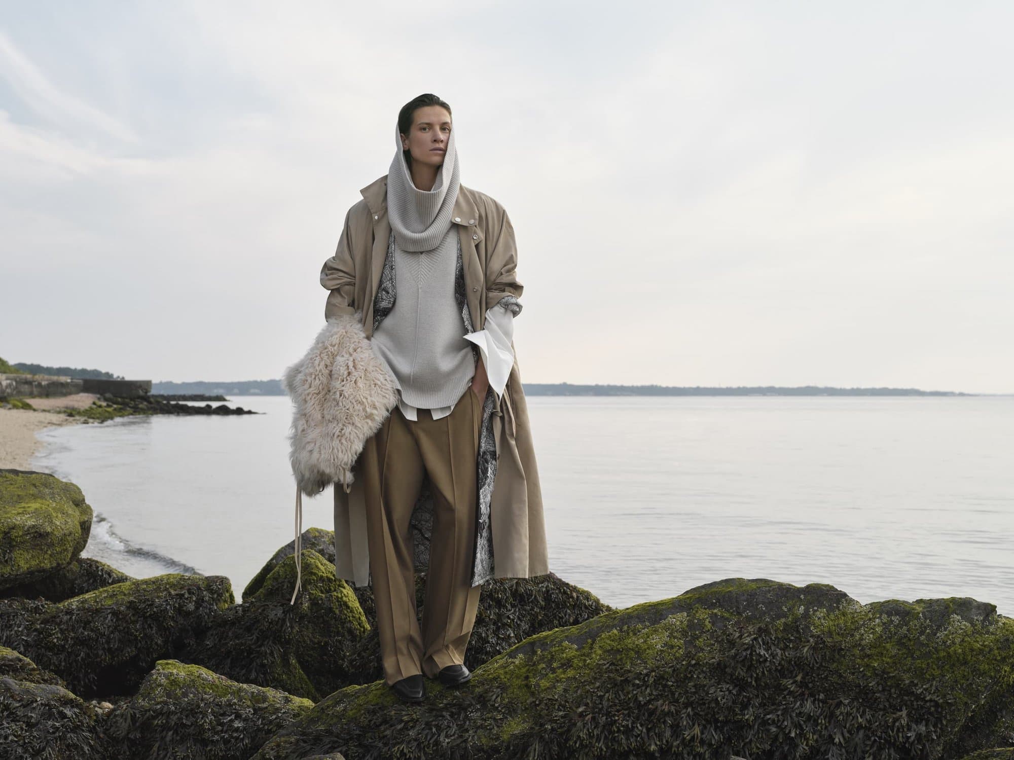 Fashion shot of a woman in a tan coat and draped scarf standing on mossy rocks near the ocean on an overcast day.