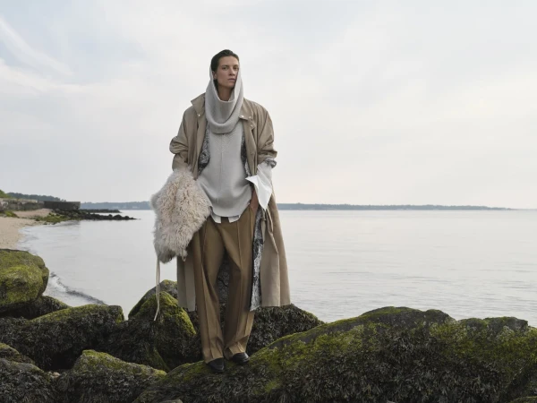 Fashion shot of a woman in a tan coat and draped scarf standing on mossy rocks near the ocean on an overcast day.