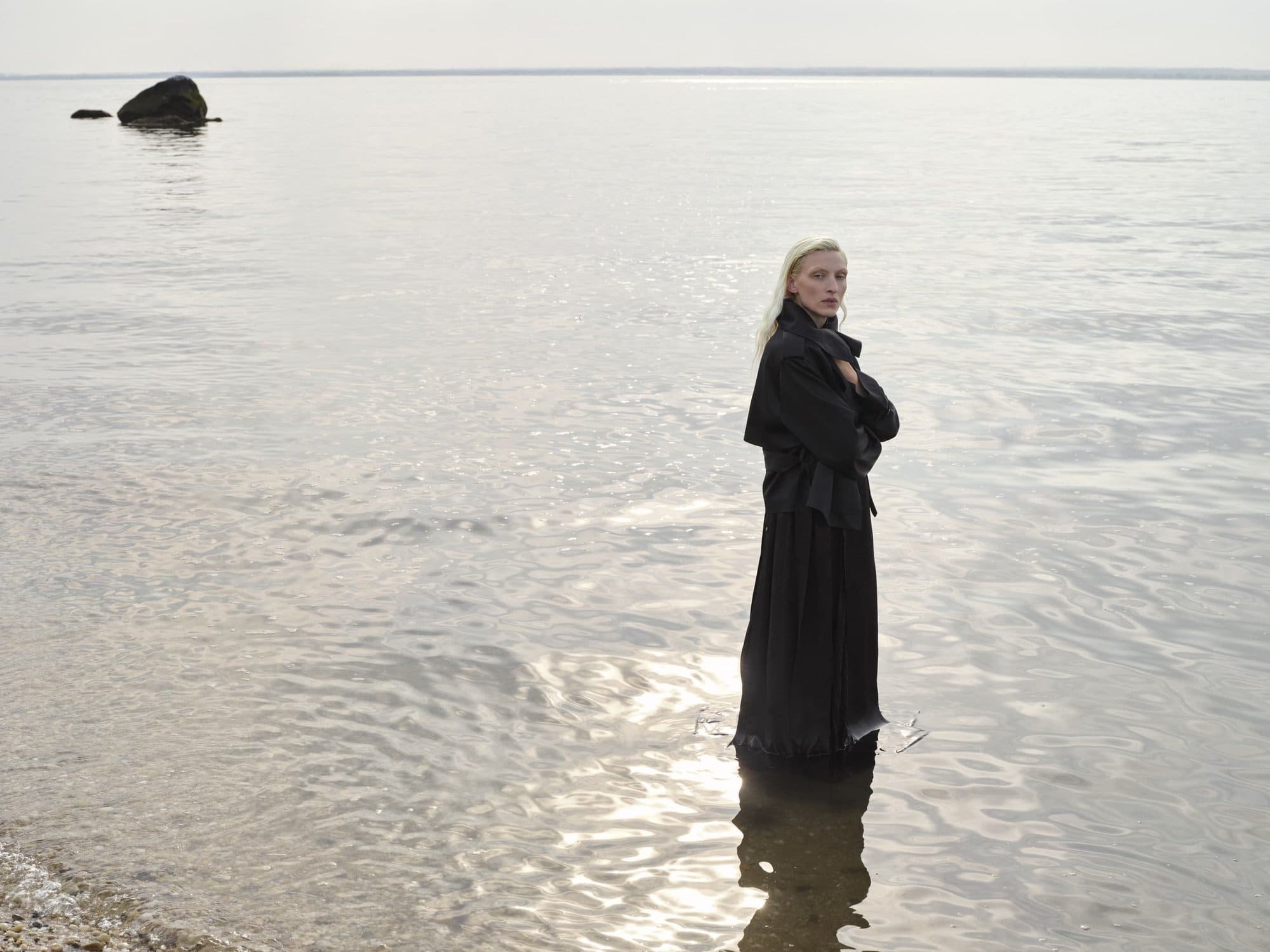 Woman stands in still water at a lakeside wearing a long black dress, her arms crossed, with a rock visible on the horizon.