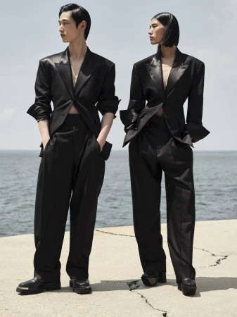 Two fashion models wearing black pantsuits stand on a stone platform with the ocean in the background under a cloudy sky.