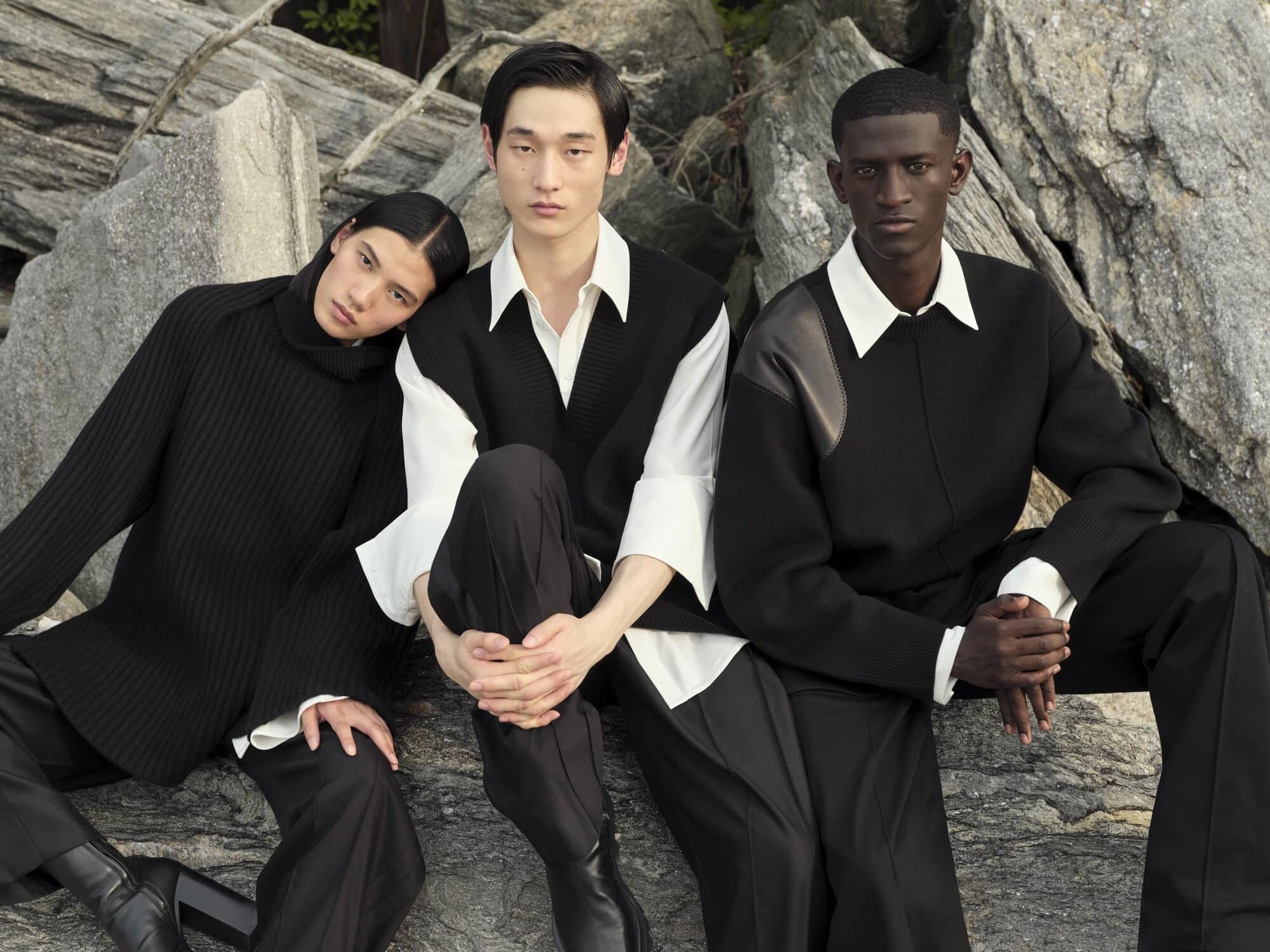 Three fashion models in coordinated black and white outfits sit on rocks in an outdoor setting, showcasing contemporary style.