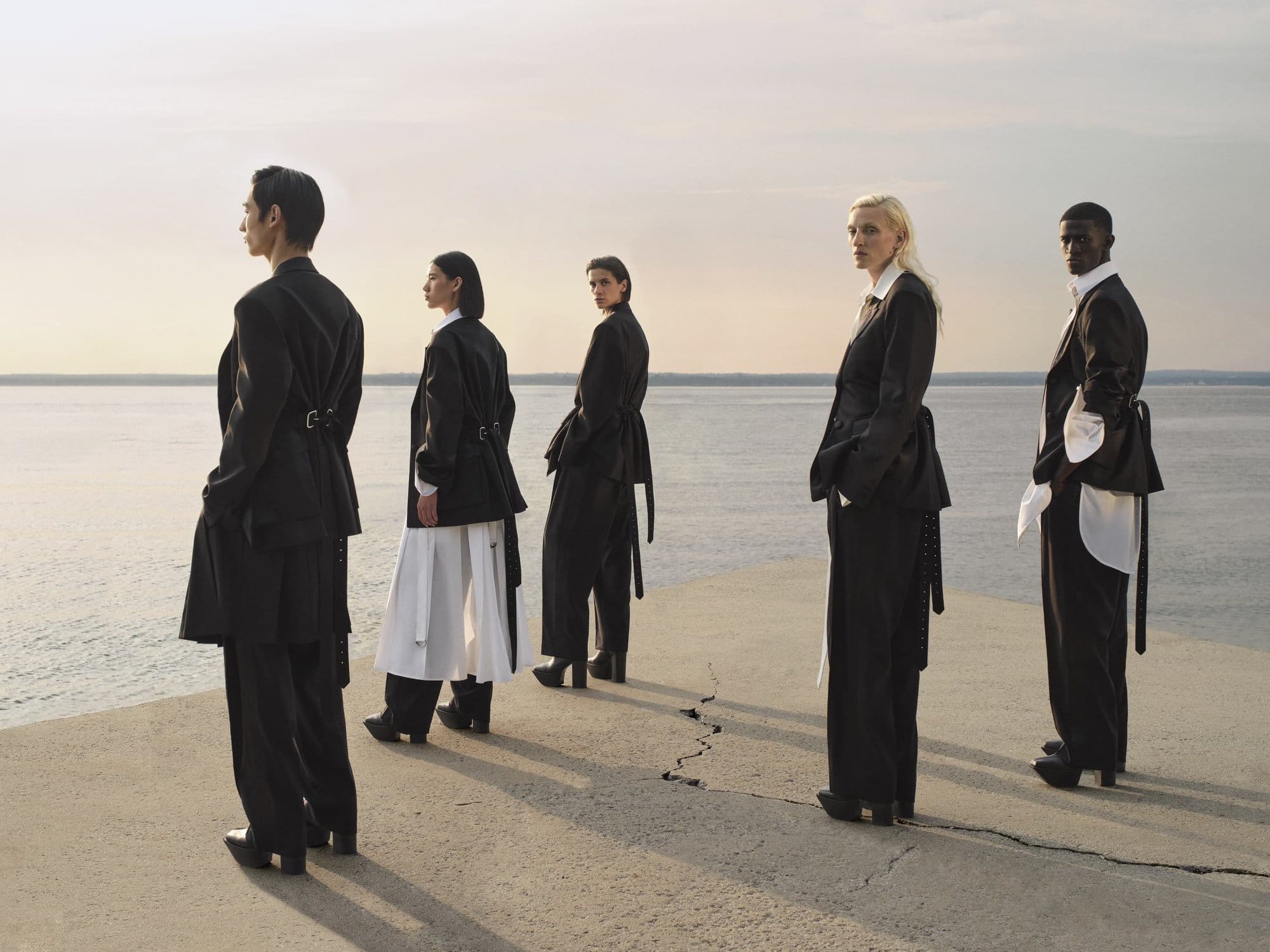 Five diverse individuals stand on a concrete platform overlooking a serene body of water, wearing dark, elegant blazers and trousers.