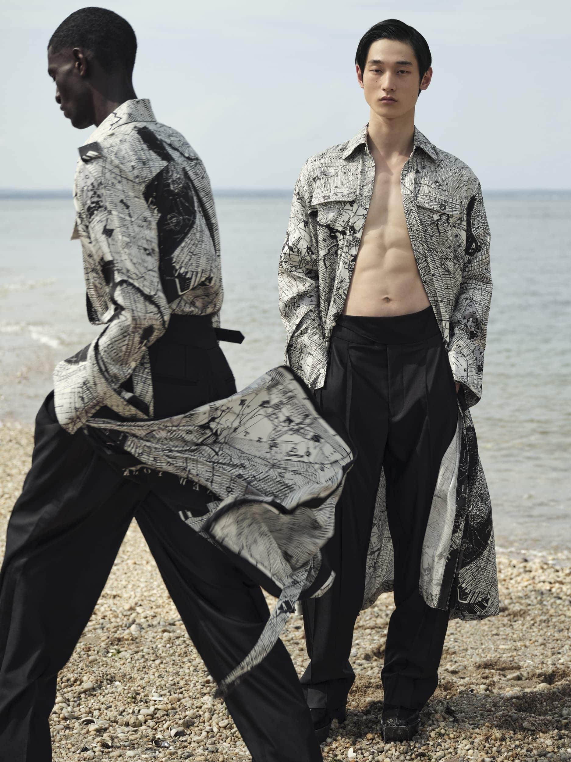 Two male fashion models in printed tops and black pants pose on a beach; one model is bare-chested in an open shirt, ocean backdrop.