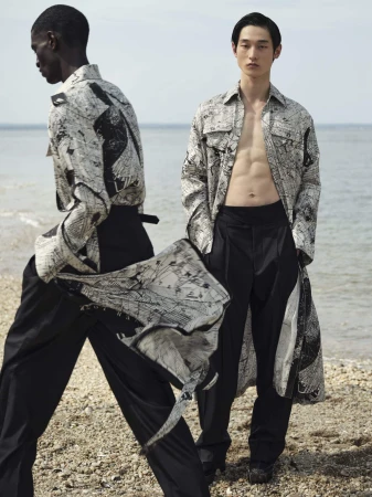 Two male fashion models in printed tops and black pants pose on a beach; one model is bare-chested in an open shirt, ocean backdrop.