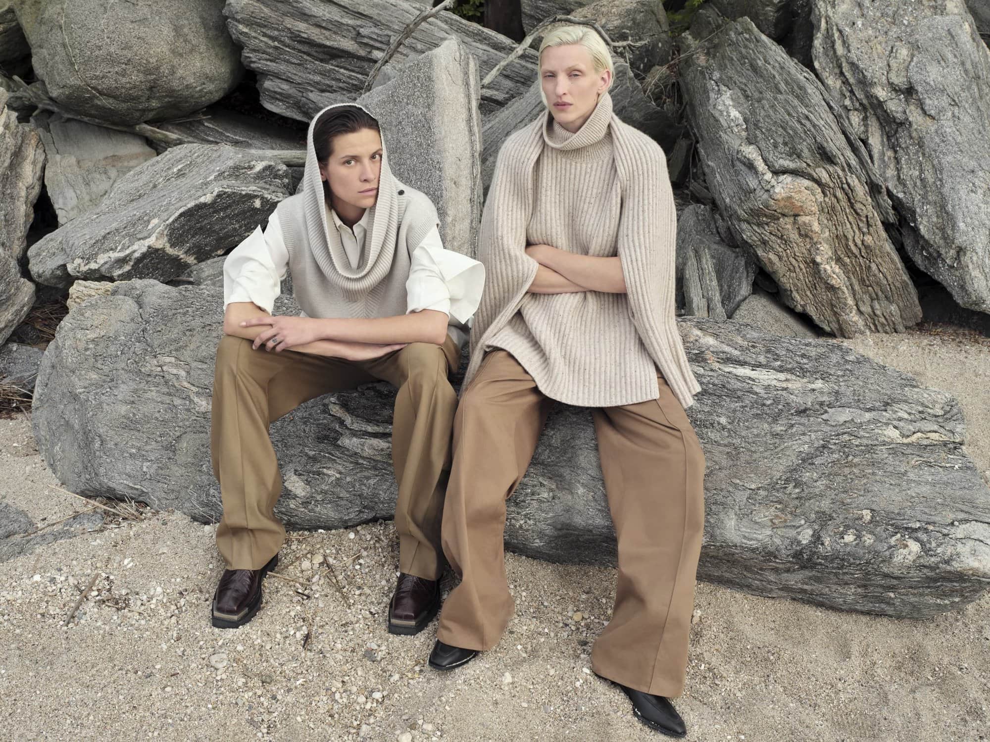 Two models pose on a rocky beach. One wears a hooded sweater vest and the other a large turtleneck knit sweater with tan wide-legged pants.