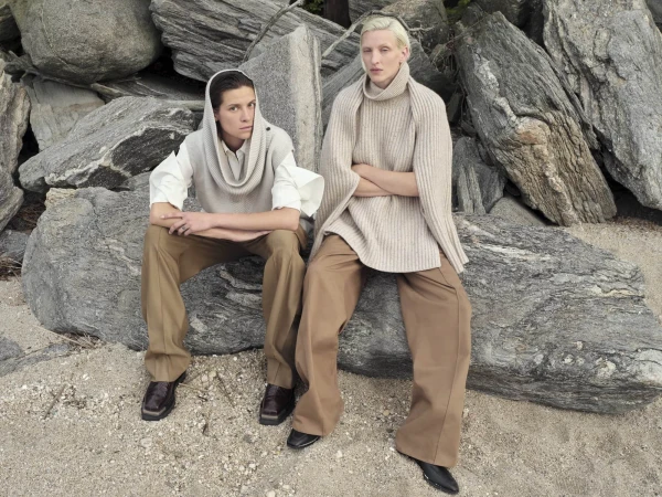 Two models pose on a rocky beach. One wears a hooded sweater vest and the other a large turtleneck knit sweater with tan wide-legged pants.