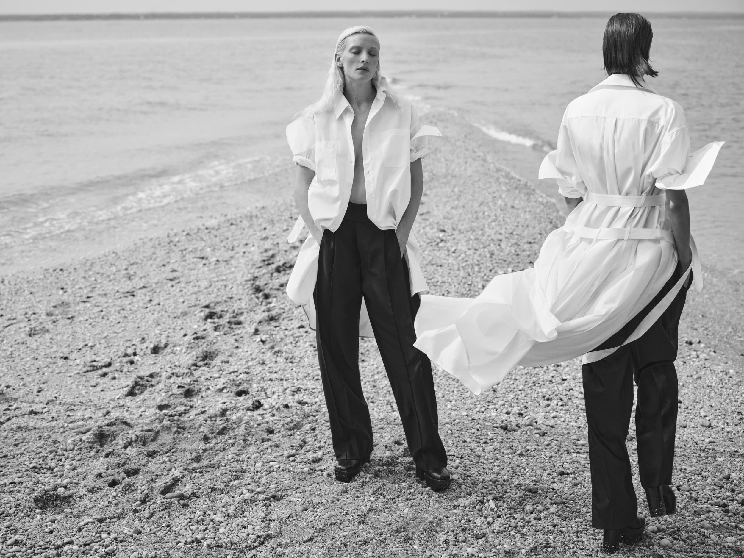 Monochrome fashion shoot on a beach featuring two models in oversized white shirts and dark trousers against a sea backdrop.