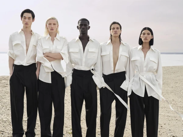 Five fashion models standing in a row on a beach wearing matching white button-down shirts and wide-leg black trousers.