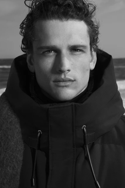 Monochrome portrait of a male model with curly hair, wearing a dark hooded coat and scarf, set against a blurred beach background.