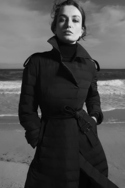 Monochrome fashion portrait of an adult female in a trench coat on a beach with soft waves and a bright sky with light clouds.