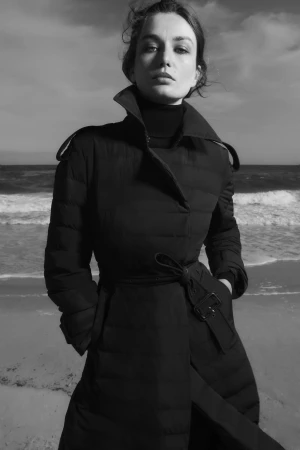 Monochrome fashion portrait of an adult female in a trench coat on a beach with soft waves and a bright sky with light clouds.