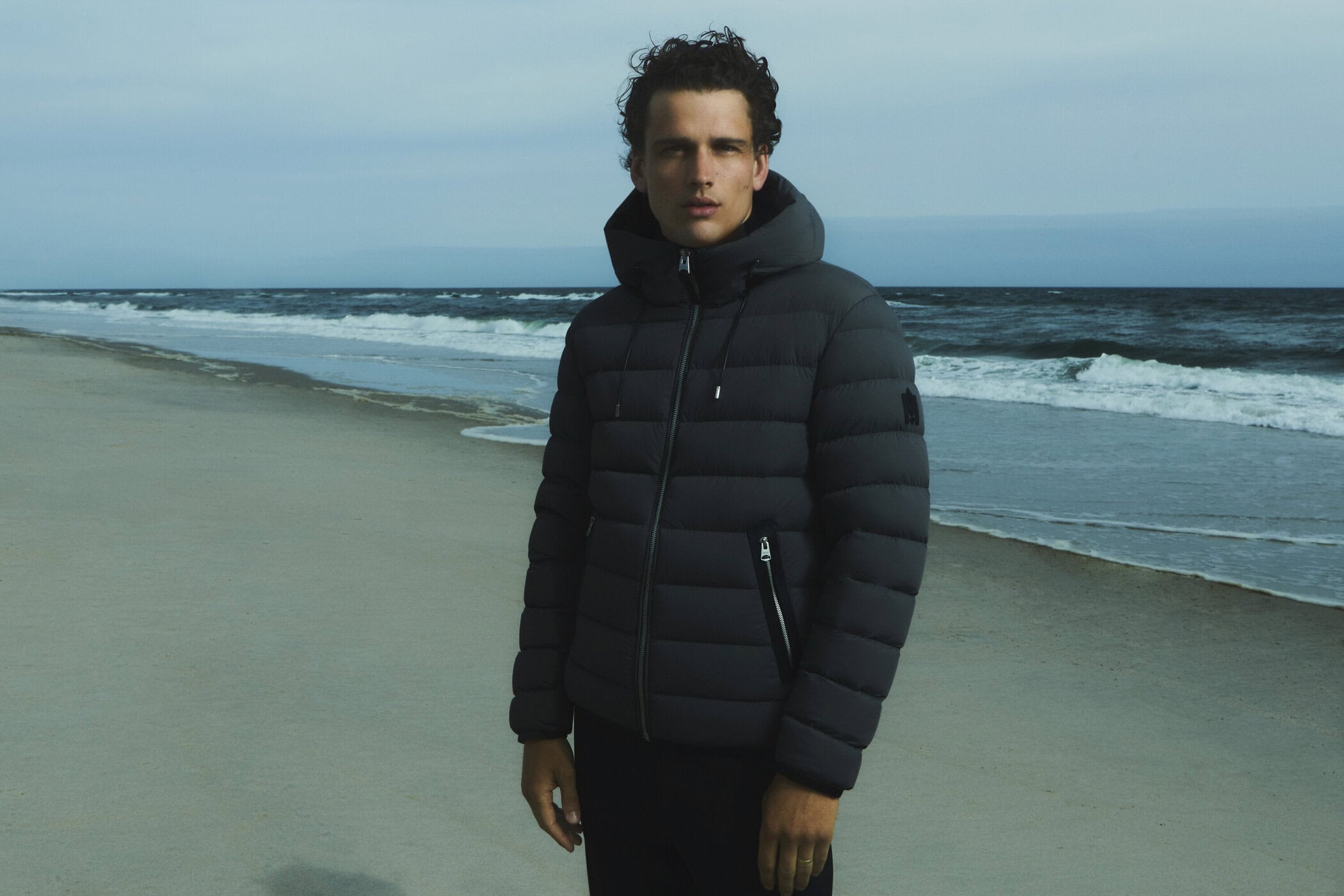 A man stands on a sandy beach in a hooded puffer jacket with ocean and cloudy sky in the background for a fashion editorial shot.