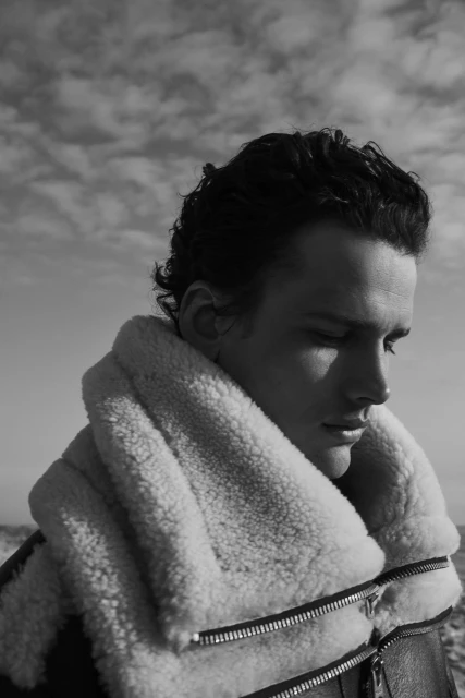 Black and white portrait of adult male in shearling jacket against sky, looking downward with soft, natural lighting.
