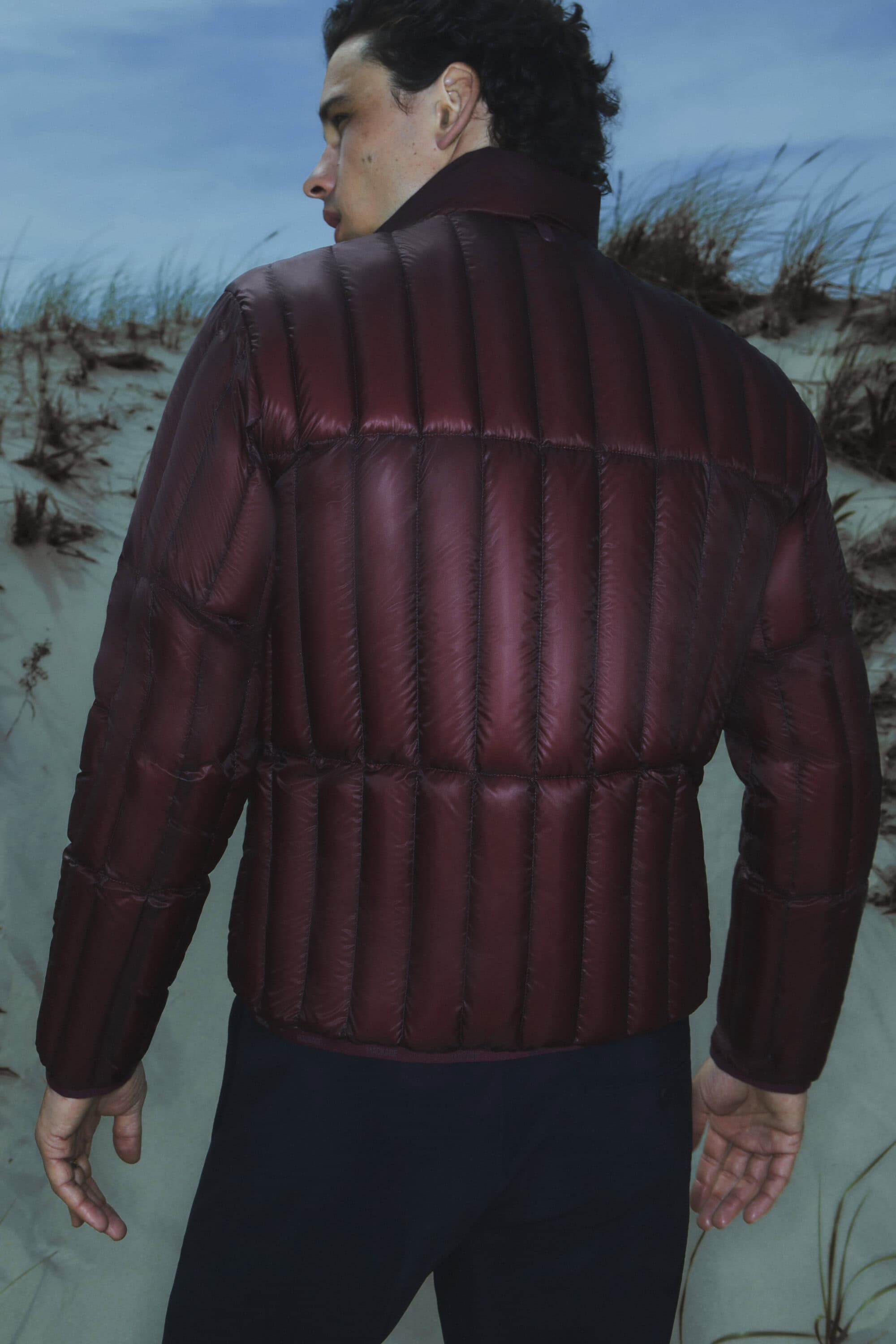 A person stands on a sandy dune with sparse vegetation wearing a burgundy puffer jacket and black pants, against a blue sky backdrop.