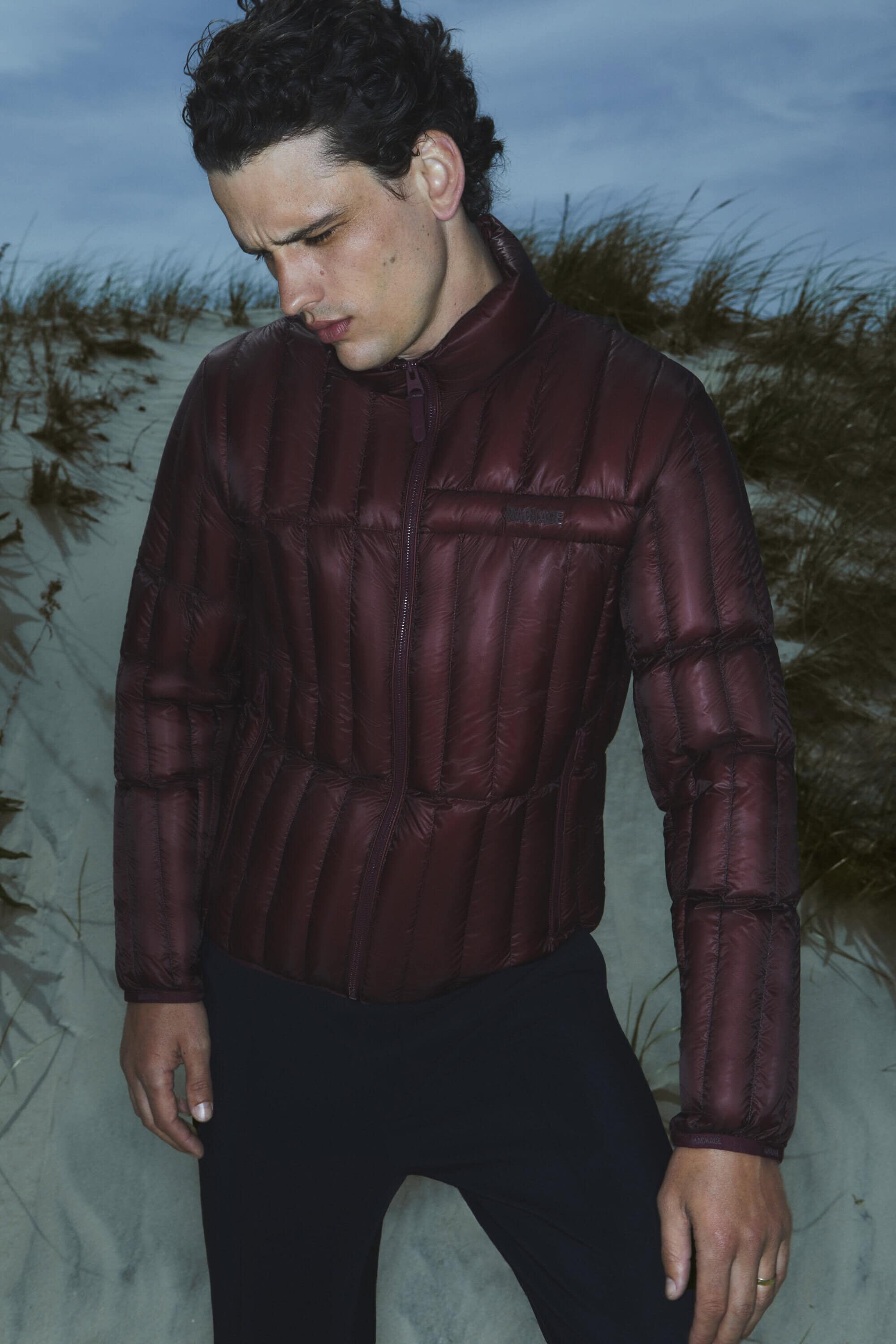 A man wearing a burgundy quilted puffer jacket stands on a sandy beach at dusk, looking down with a somber expression.