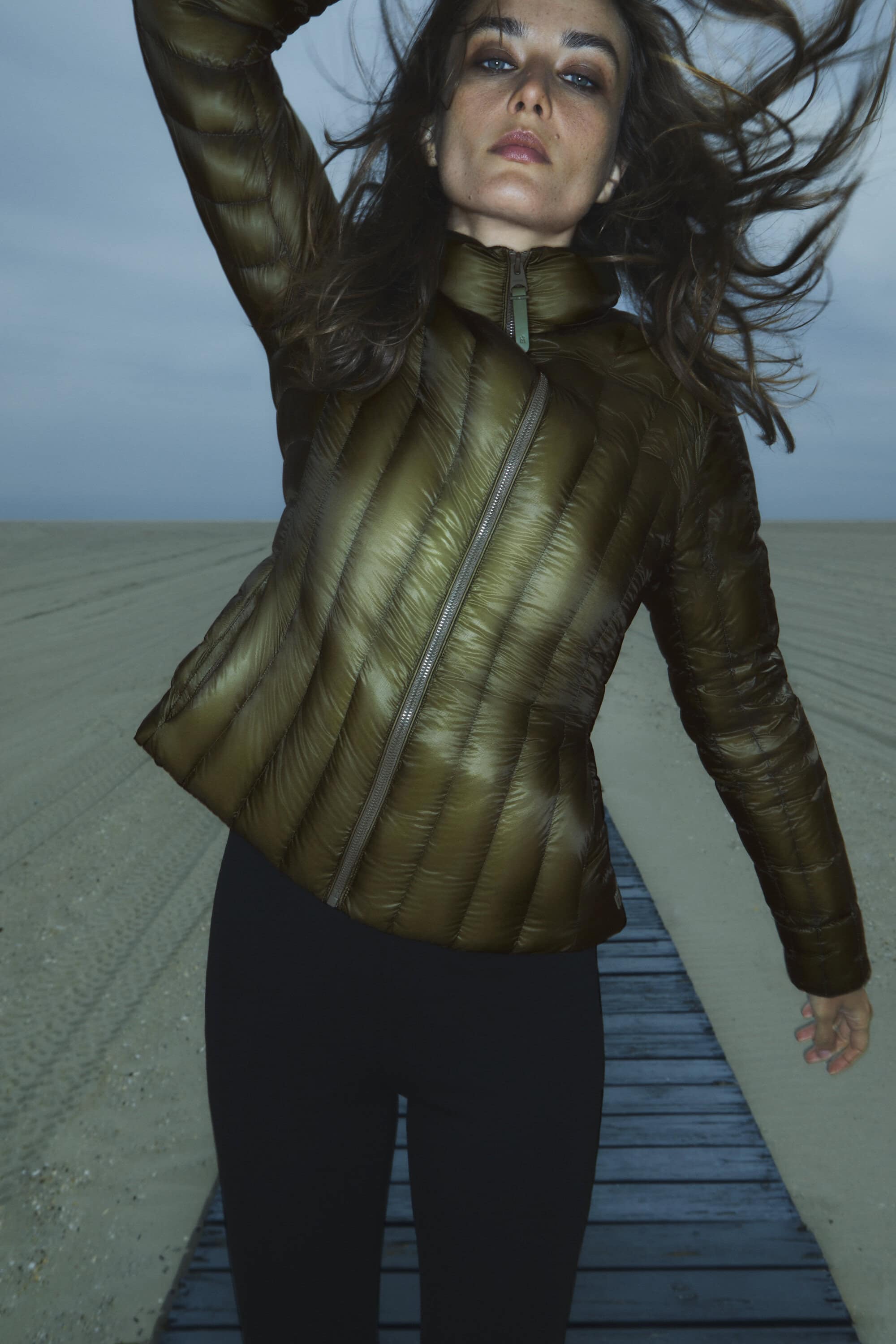 A woman in a quilted jacket poses on a beach boardwalk under an evening sky, showcasing fashion and outdoor lifestyle photography.