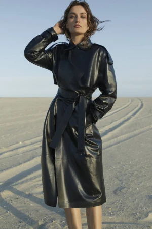 Model wearing a glossy black leather belted coat standing on a sandy beach under a clear sky.