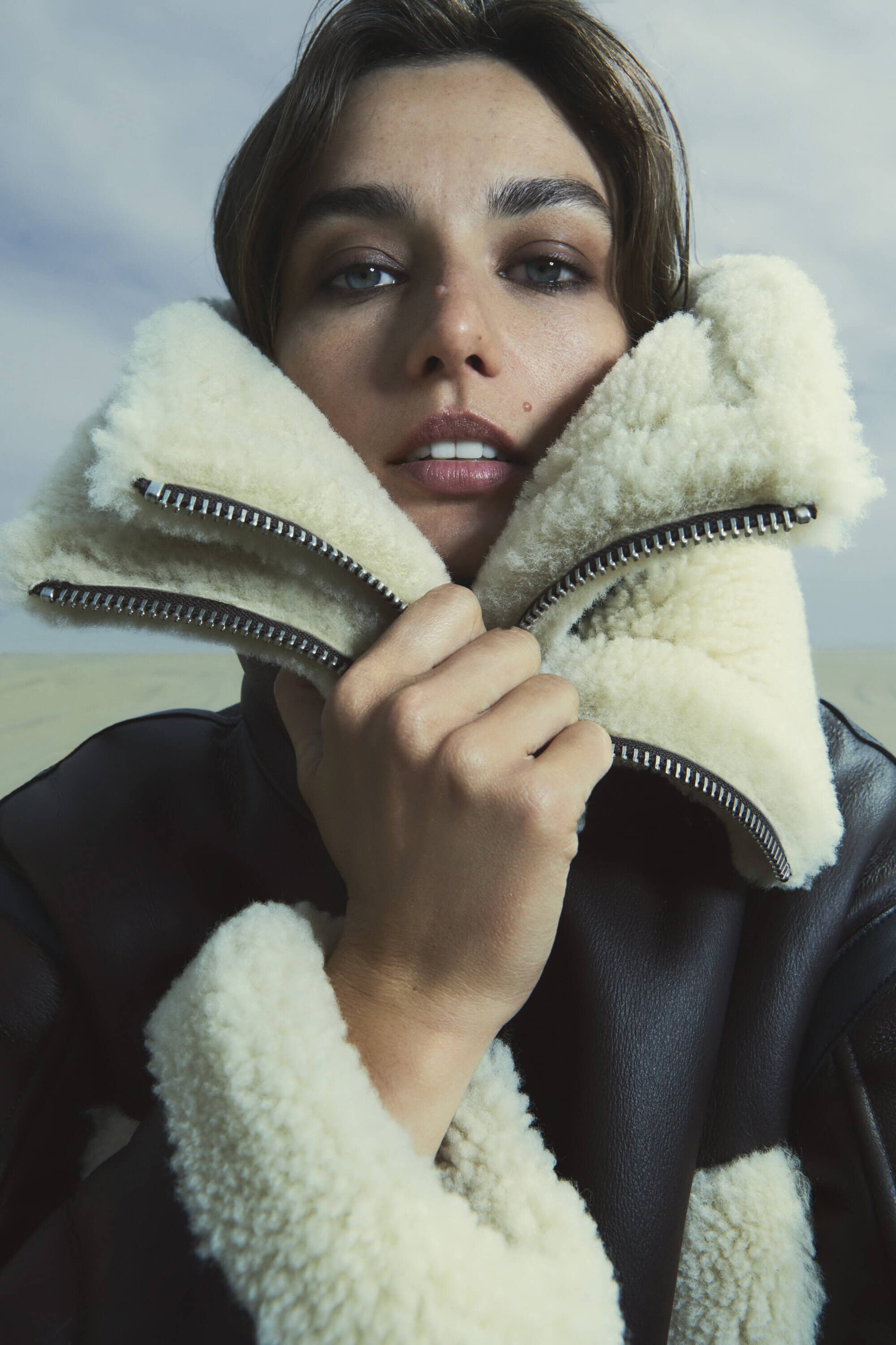 Close-up portrait of a brunette adult female wearing a brown shearling bomber jacket against a blurred sky background.