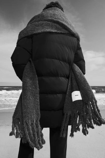 Back view of a person in a black puffer jacket and gray scarf standing on a sandy beach with the ocean and sky in the background.