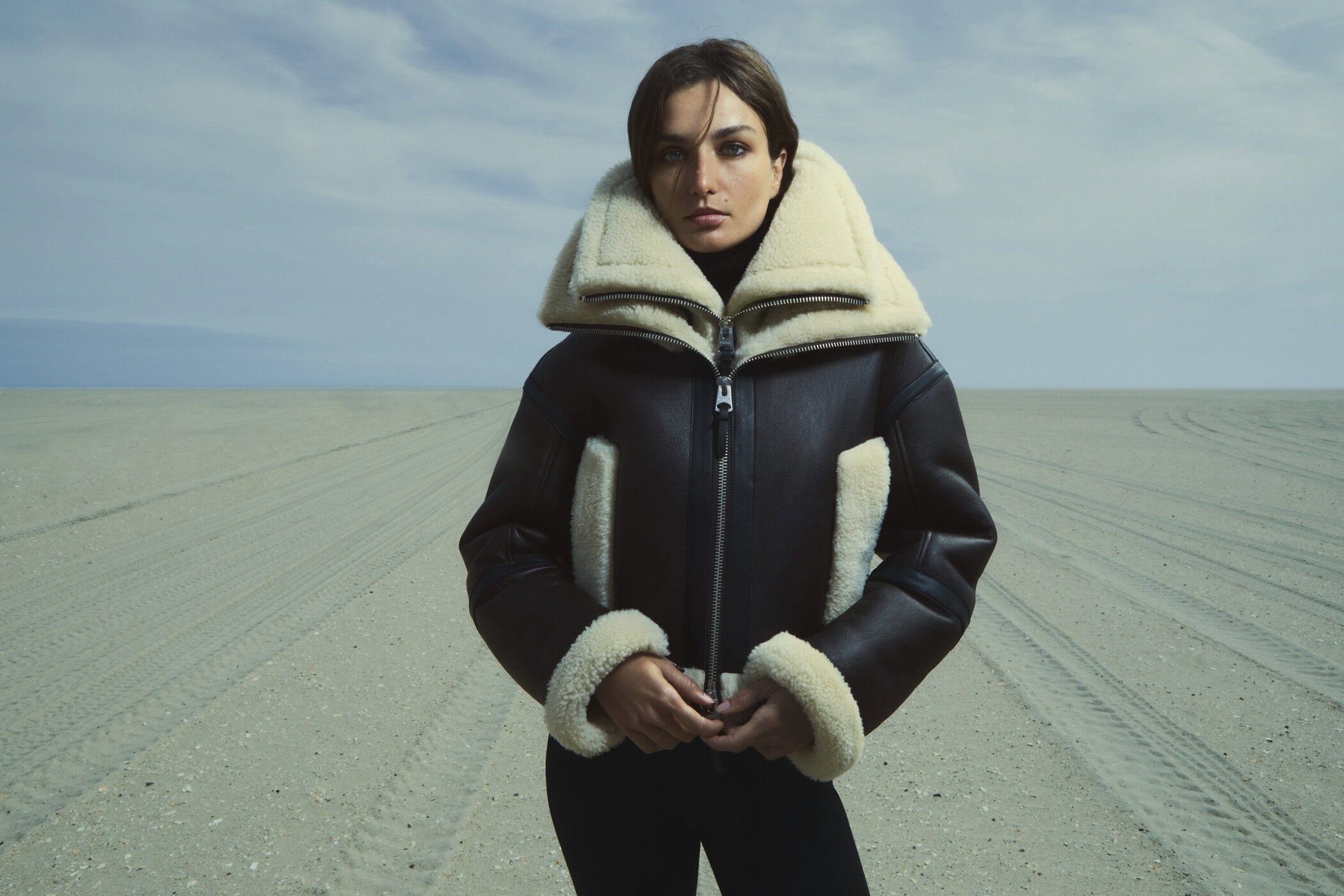 Woman in shearling-lined jacket on beach. Close-up portrait of female model in fashionable outerwear. Neutral tones. Fashion editorial shot.
