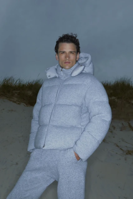 Adult male poses wearing a light gray puffer jacket and matching sweatpants in a beach environment with tall grasses and an overcast sky.