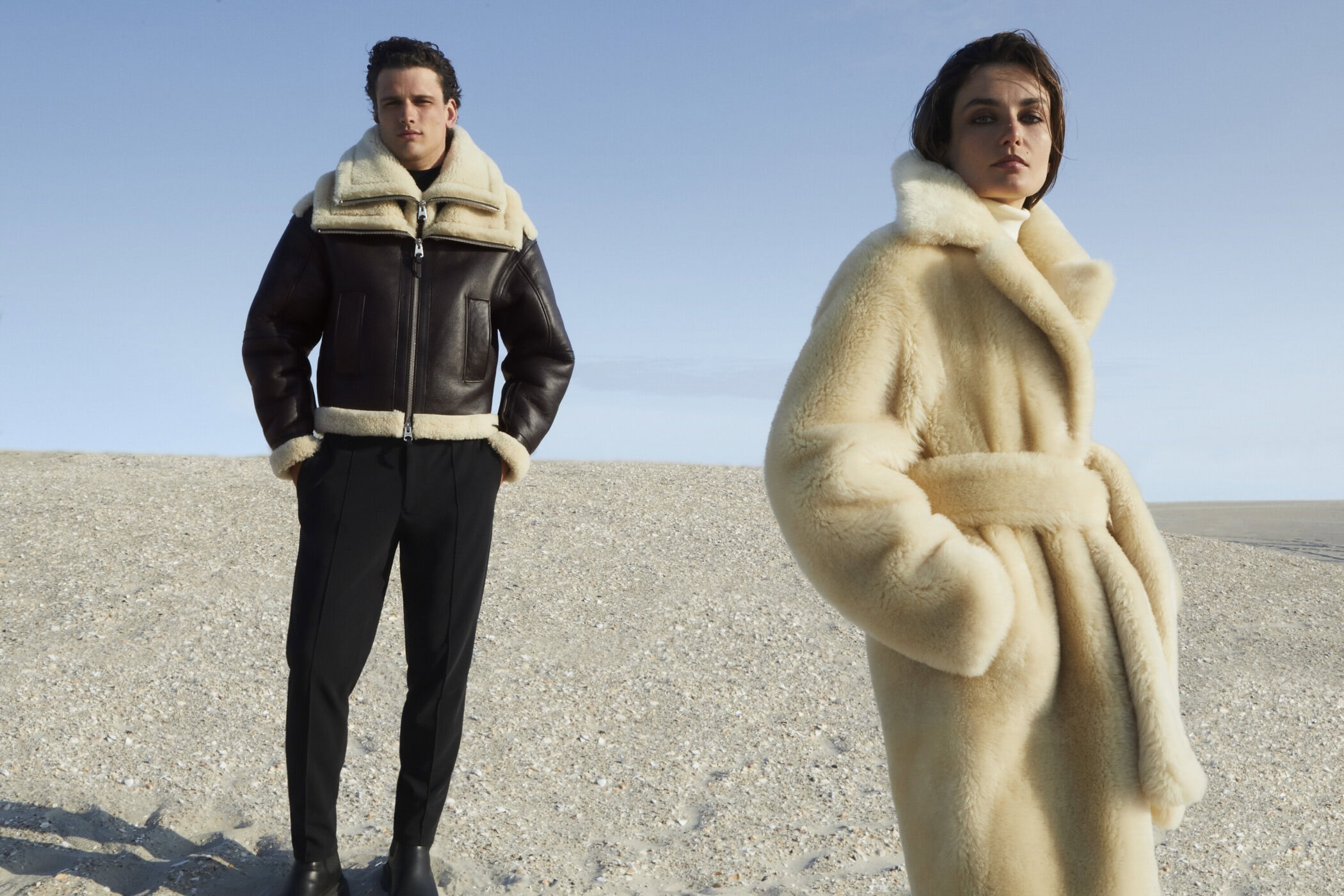 Two fashion models in shearling coats on a sand dune under a clear sky. One wears dark brown, the other cream-colored.