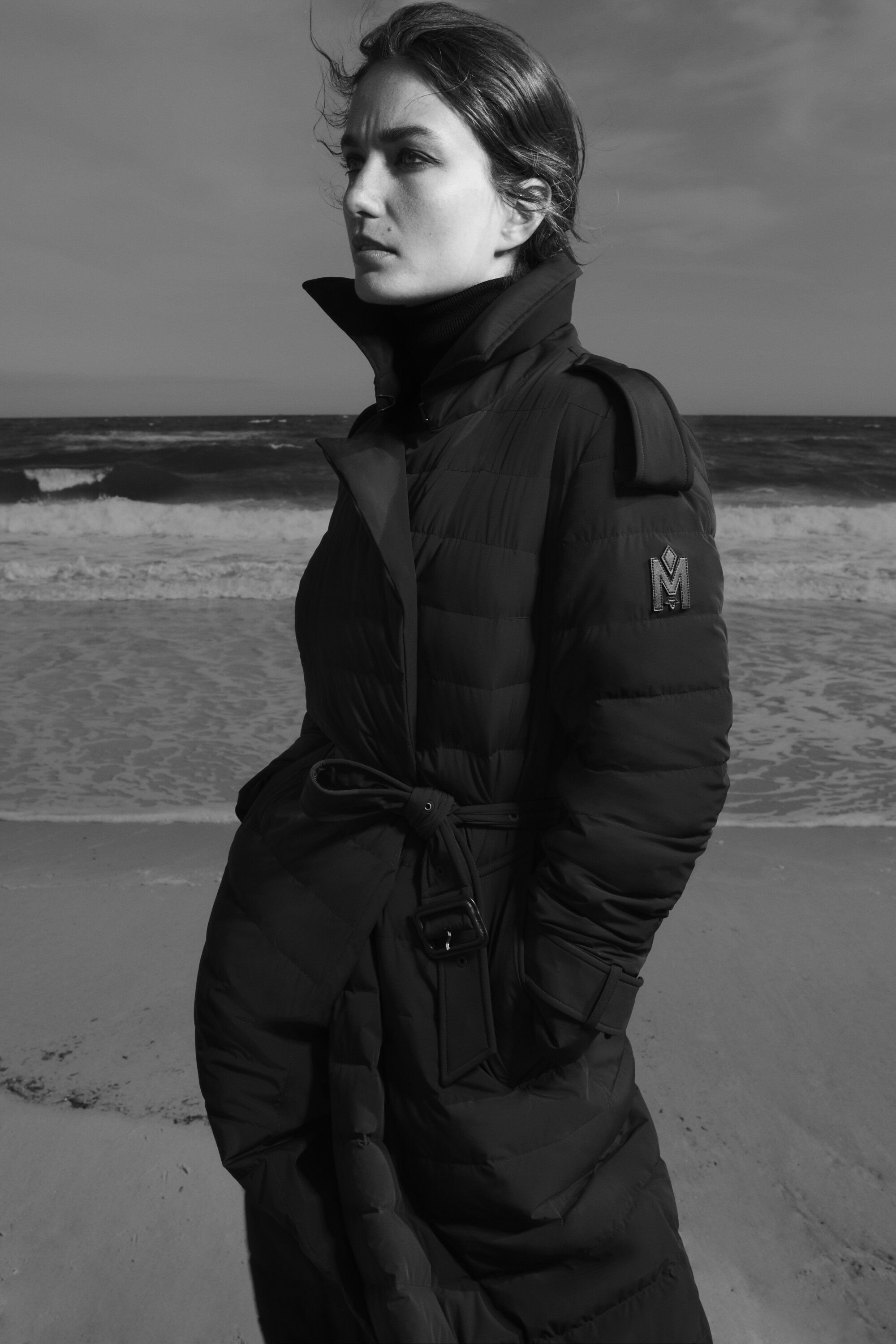 Monochrome portrait of a woman in a black puffer coat standing on a sandy beach with ocean waves in the background under a cloudy sky.
