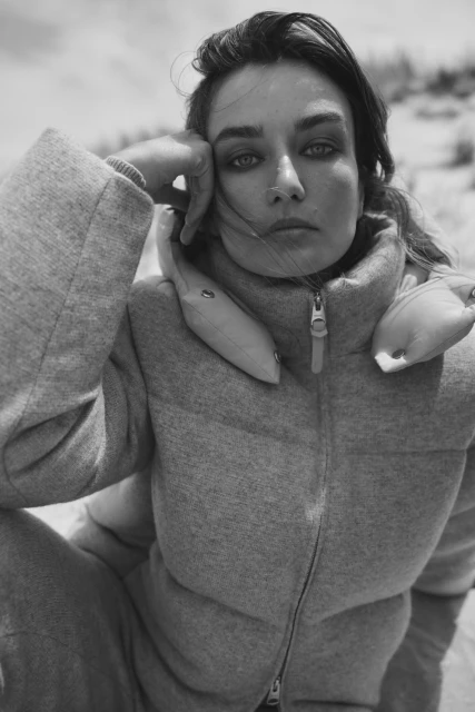 Black and white portrait of a woman on a beach, wearing a puffy coat. The camera captures her head and shoulders.