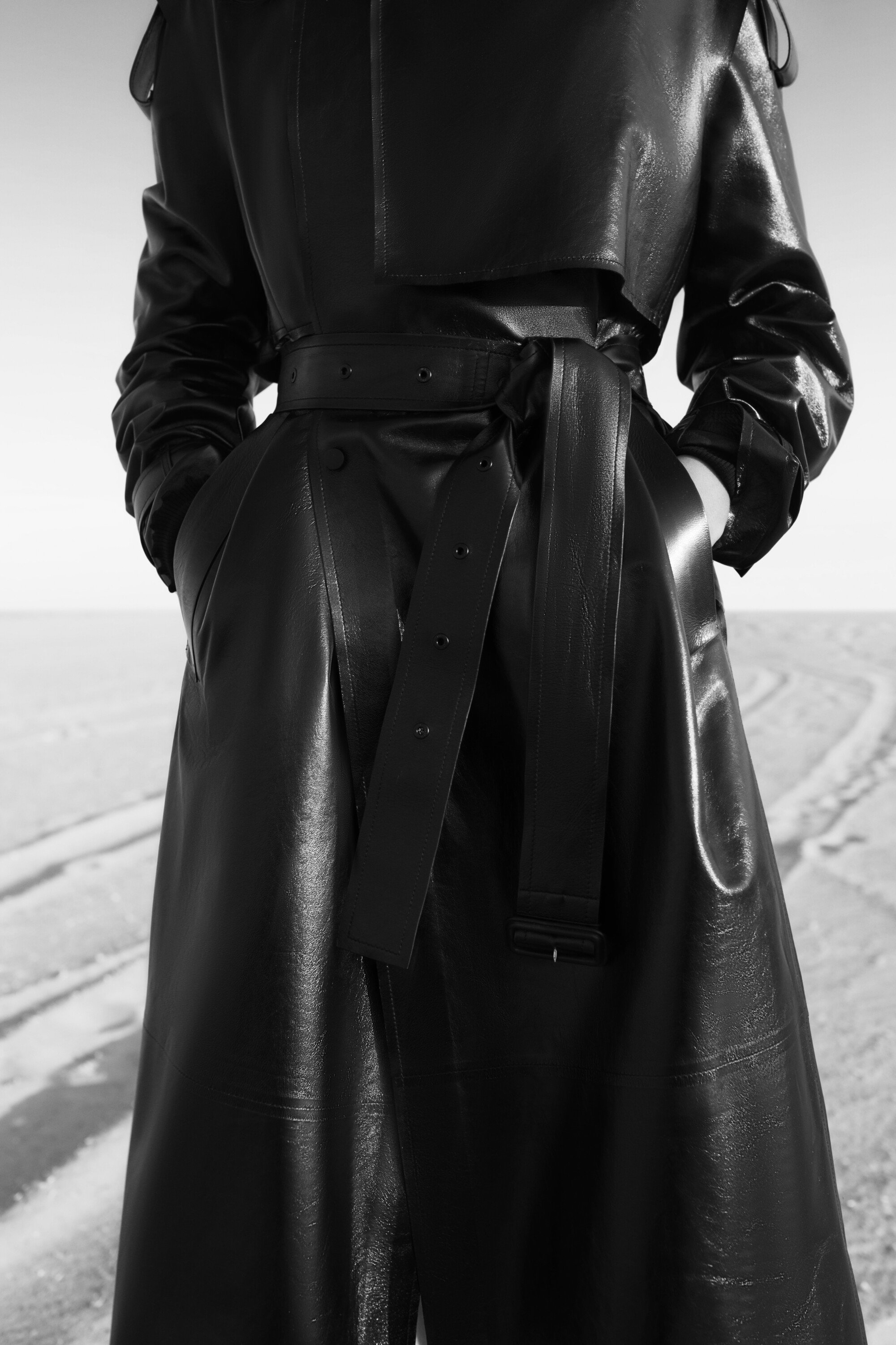Close-up of a person wearing a long, belted, shiny black trench coat, standing outdoors in a bright, neutral-toned environment.
