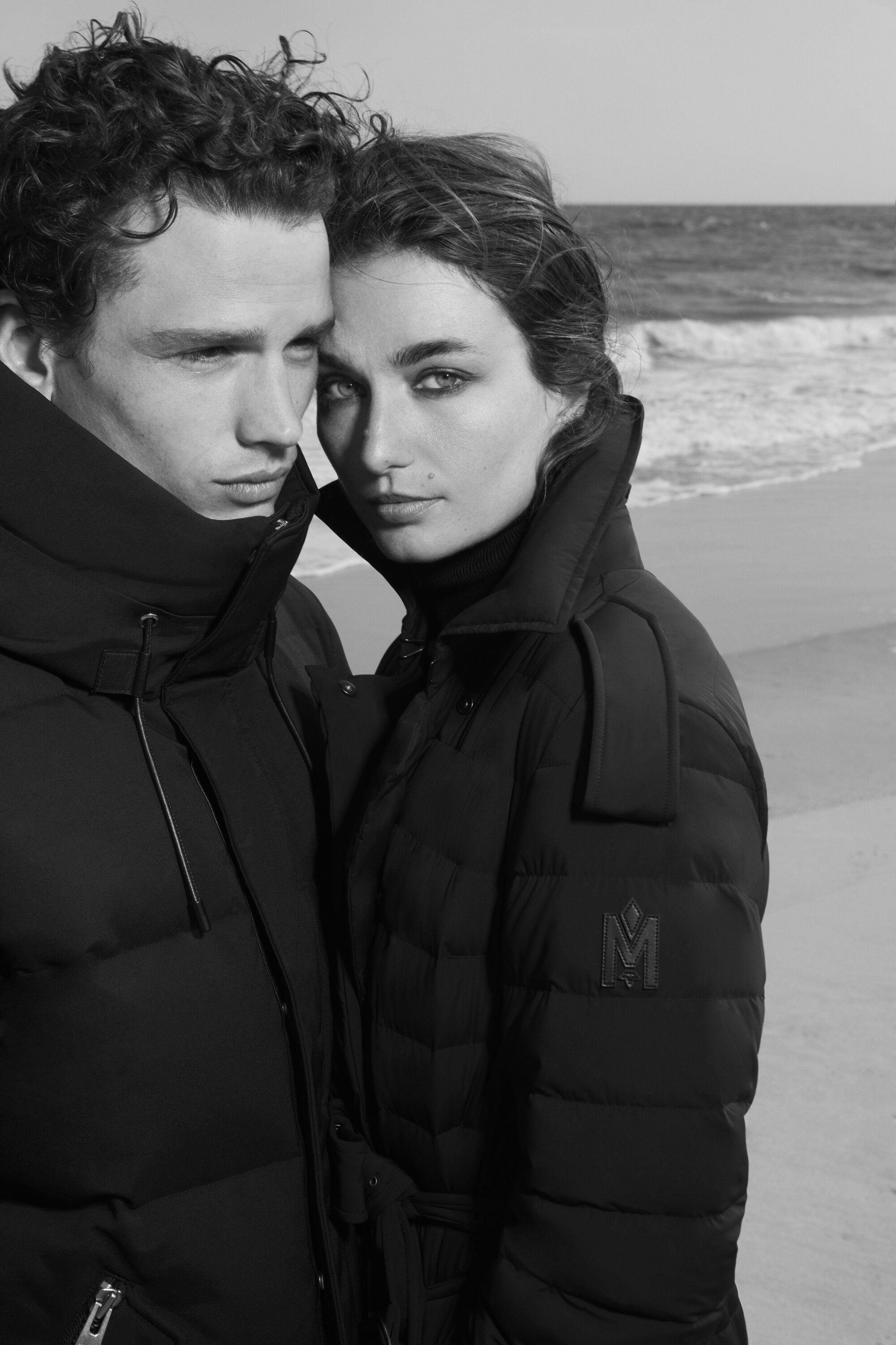 A couple in black jackets pose on a beach with ocean waves. Black and white fashion shot emphasizing winter outerwear. Focus on cold weather apparel.