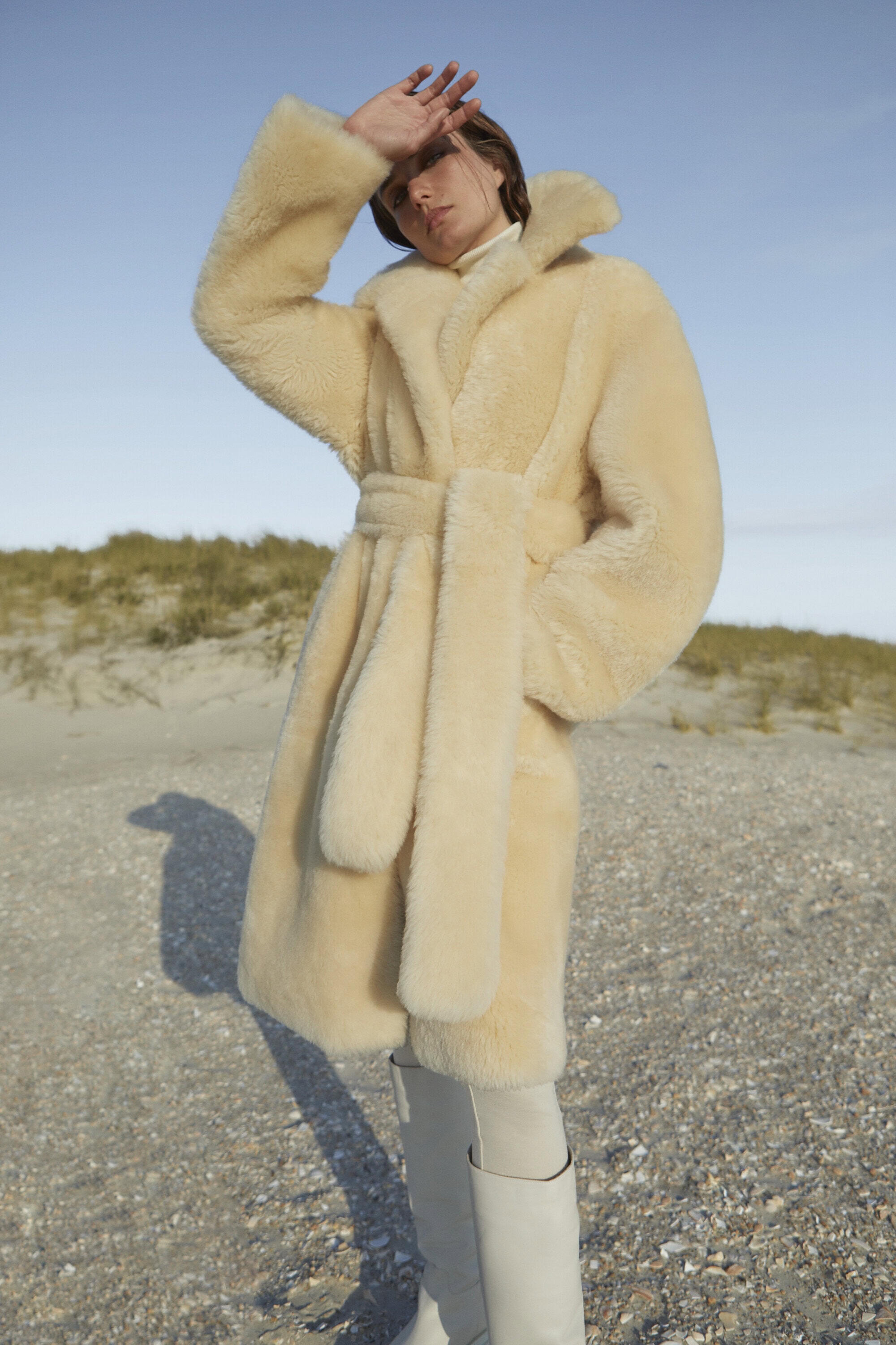 A woman in a cream coat poses on a beach, shielding her eyes from the sun against a clear sky, wearing cream knee-high boots.