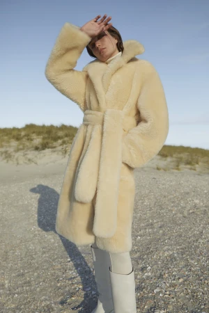 A woman in a cream coat poses on a beach, shielding her eyes from the sun against a clear sky, wearing cream knee-high boots.