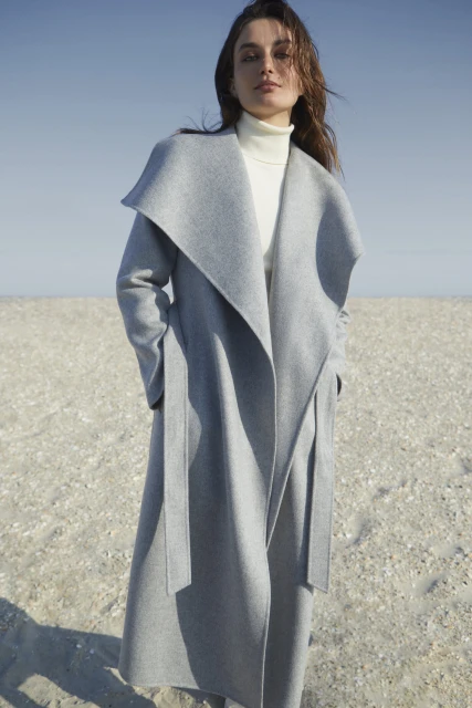 A woman models a light gray wool coat on a beach, paired with a white turtleneck. The coat features a wide collar and belted waist.