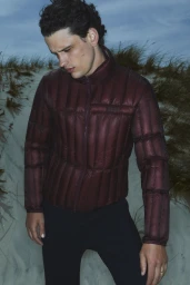 Model Simon Nessman wearing a burgundy quilted puffer jacket standing in coastal grassy dunes.