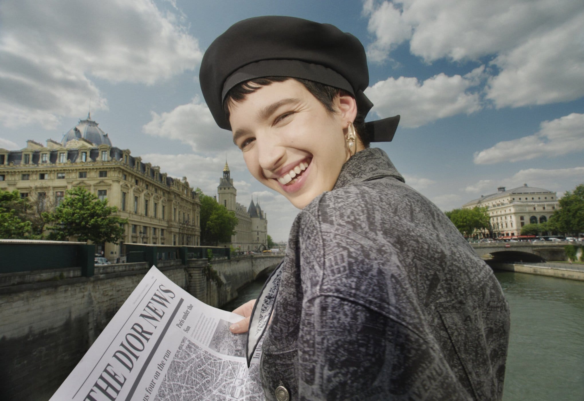 Smiling woman in a black beret reading a Dior newspaper along the Seine River in Paris, with historic buildings and a cloudy sky.