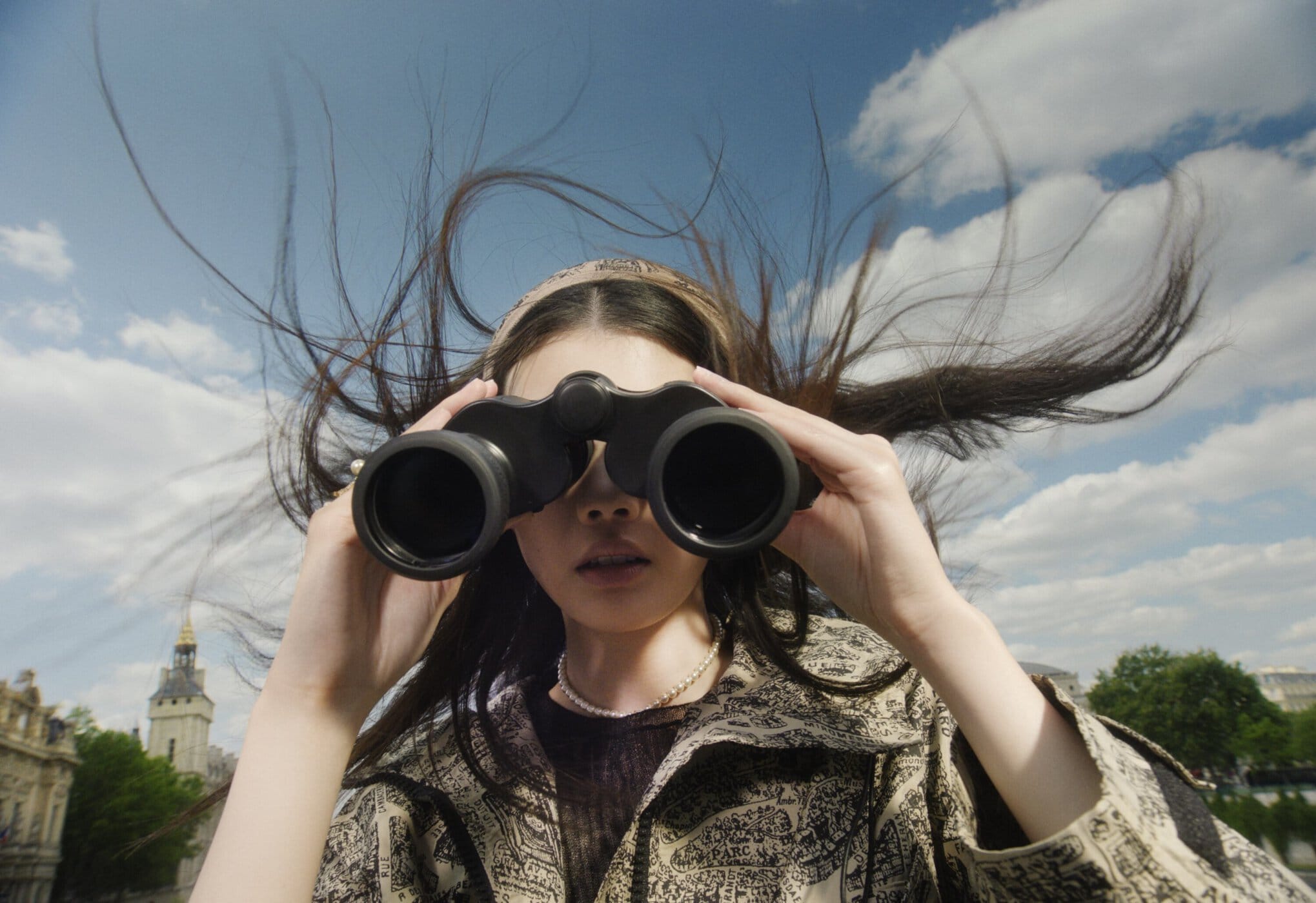 Woman in patterned coat holds binoculars against a blue sky with city buildings in the background, wind blowing her hair.