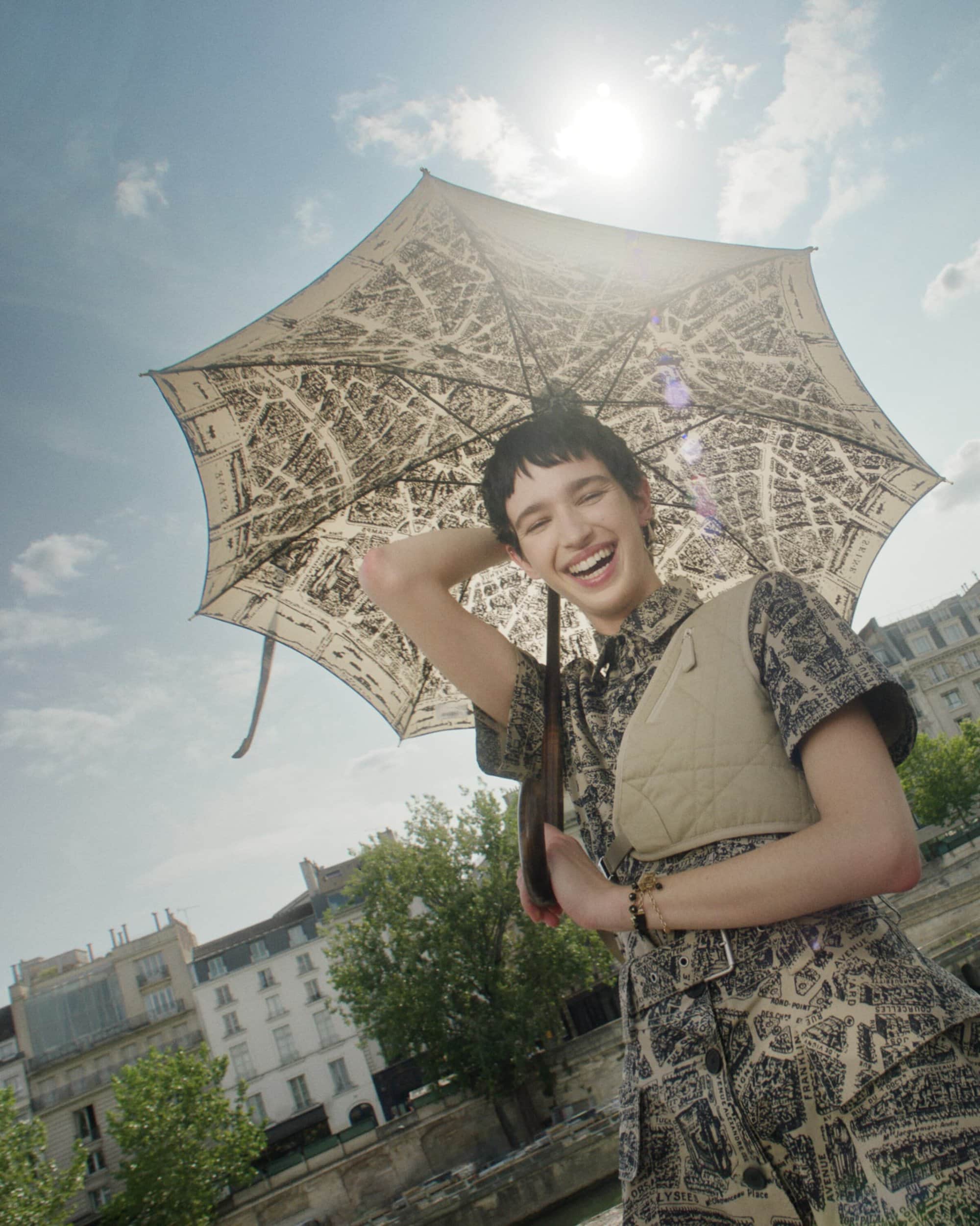 A smiling woman in patterned clothing holds a printed umbrella outdoors on a sunny day near buildings and trees, creating a joyful portrait.