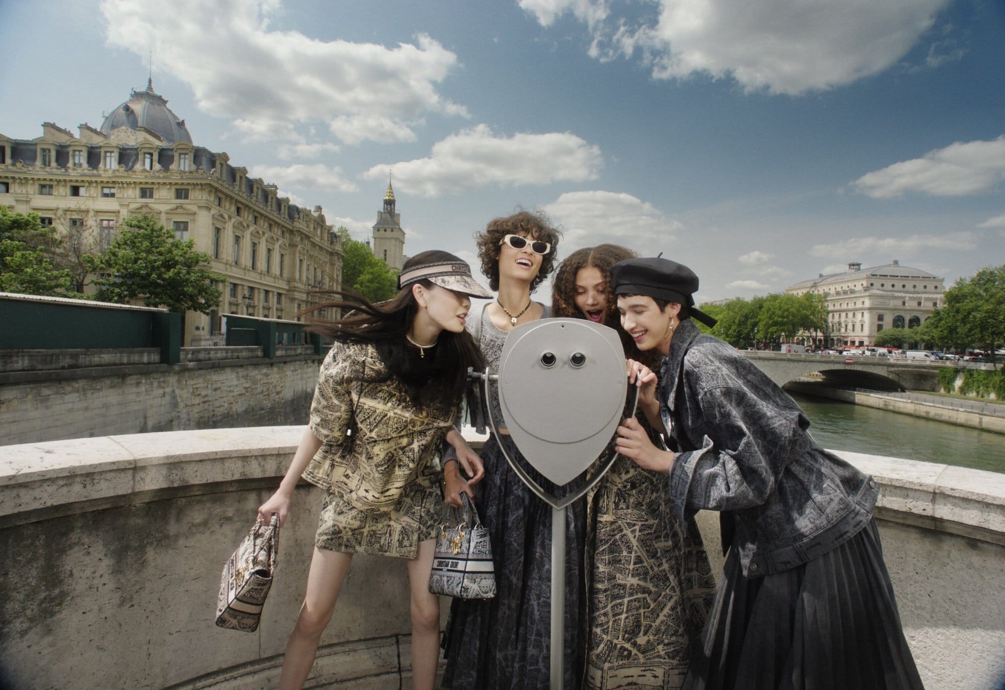 Four female models laughing together around a public view finder on a stone bridge in Paris with classic buildings and a river.
