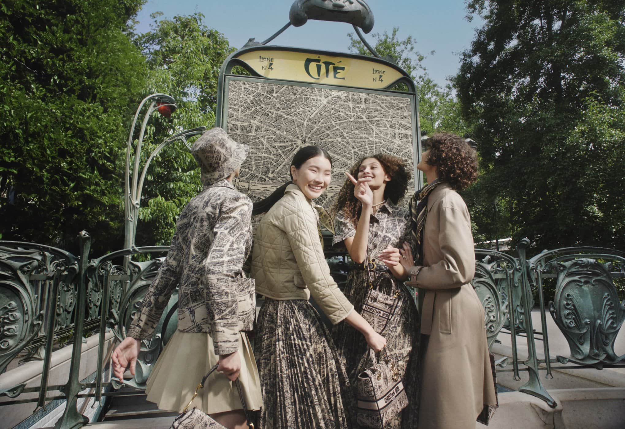 Four women model a fashion collection at a Paris Metro entrance with map backdrop, showcasing dresses, jackets, and handbags in daylight.