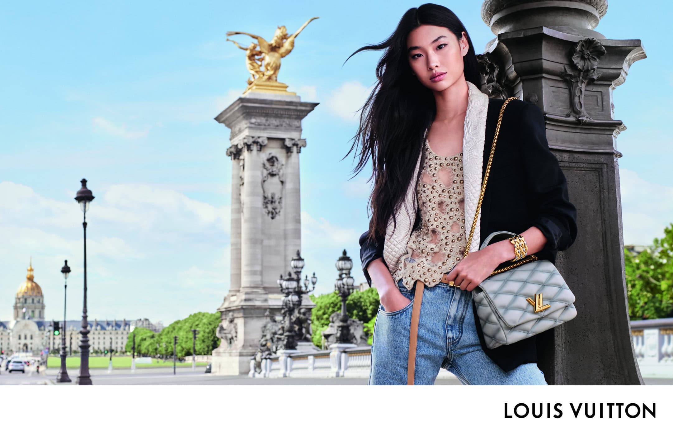 Fashion shot featuring a woman with a purse in Paris. She is wearing a jacket, top, jeans, and jewelry, with a landmark behind her.