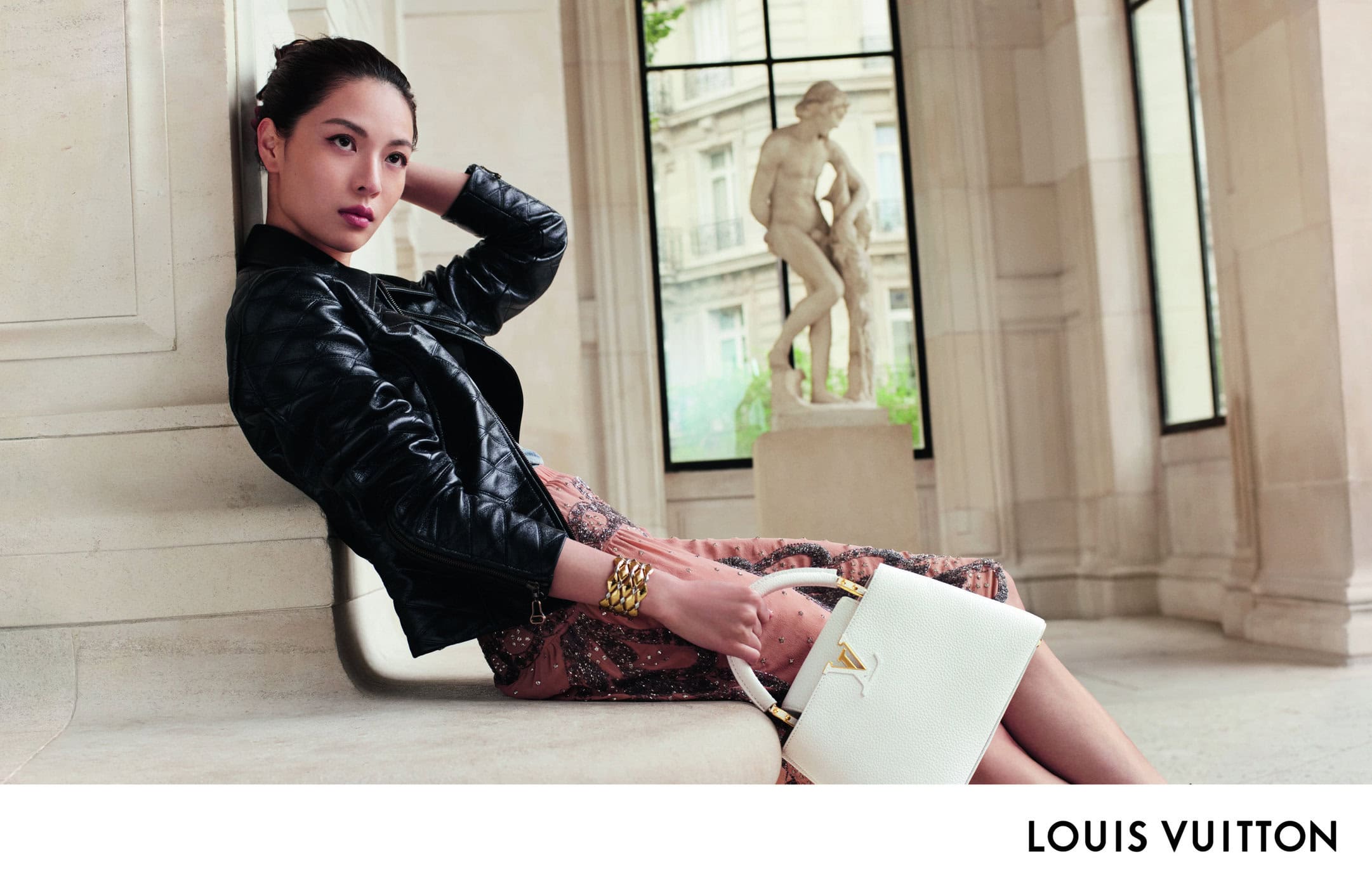 An Asian woman poses in an indoor environment near a window and sculpture, wearing a black jacket and holding a Louis Vuitton handbag.