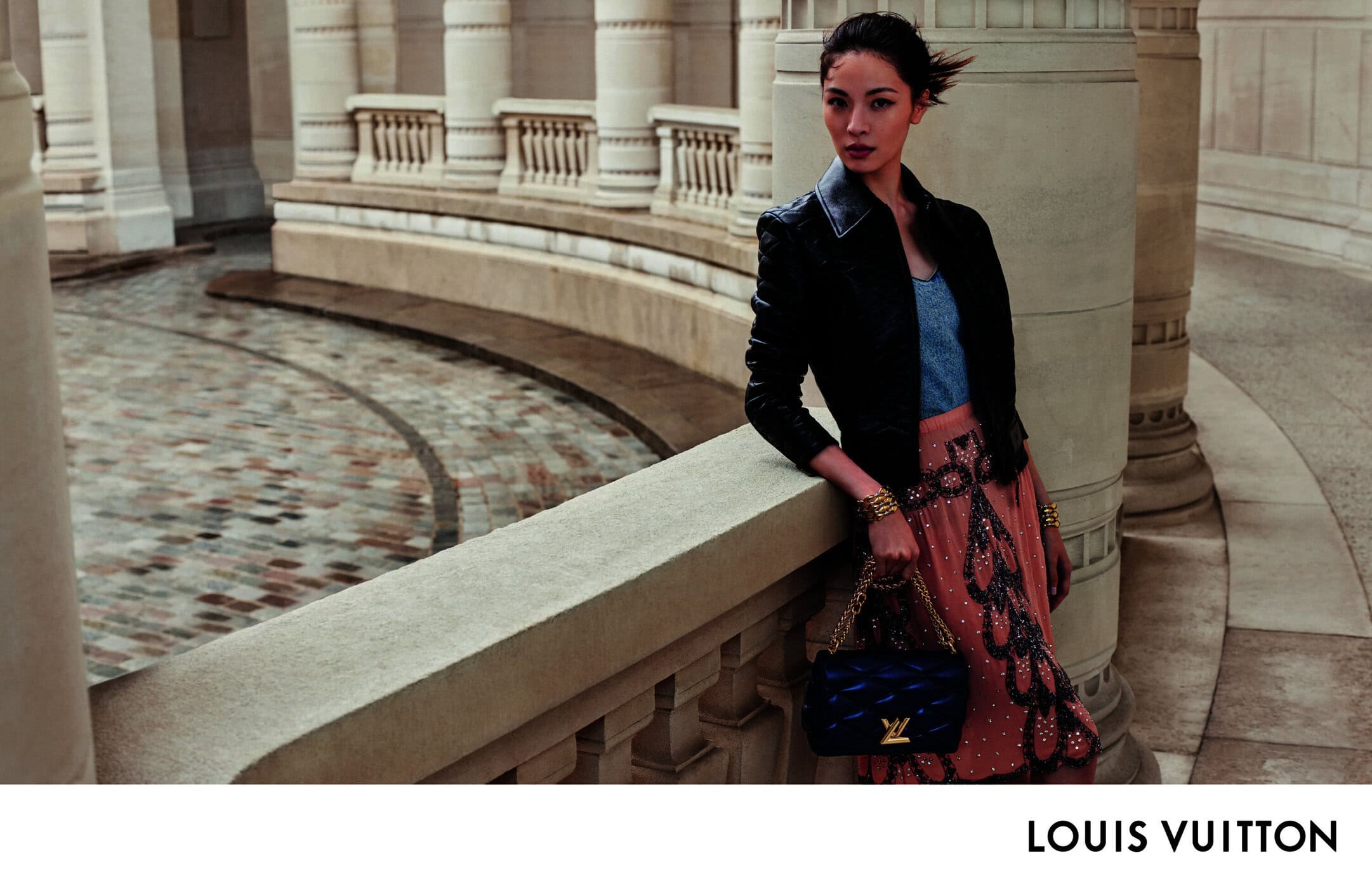 Elegant woman in black jacket and patterned skirt leans against stone balustrade, holding a blue quilted purse in a stylish outdoor fashion shot.