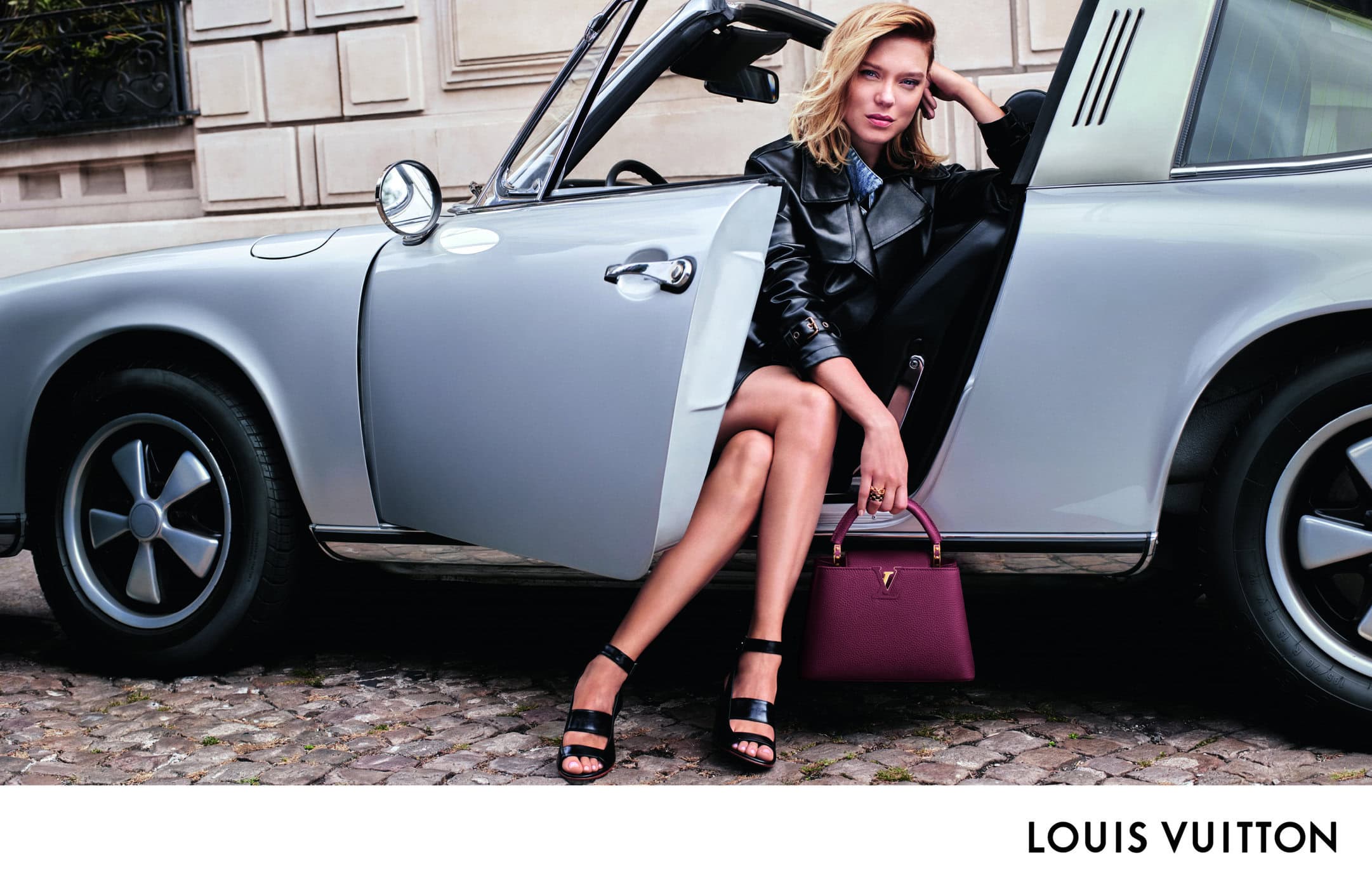 A stylish adult female model sits in a silver convertible car with a maroon Louis Vuitton handbag, wearing a black leather jacket and black sandals.
