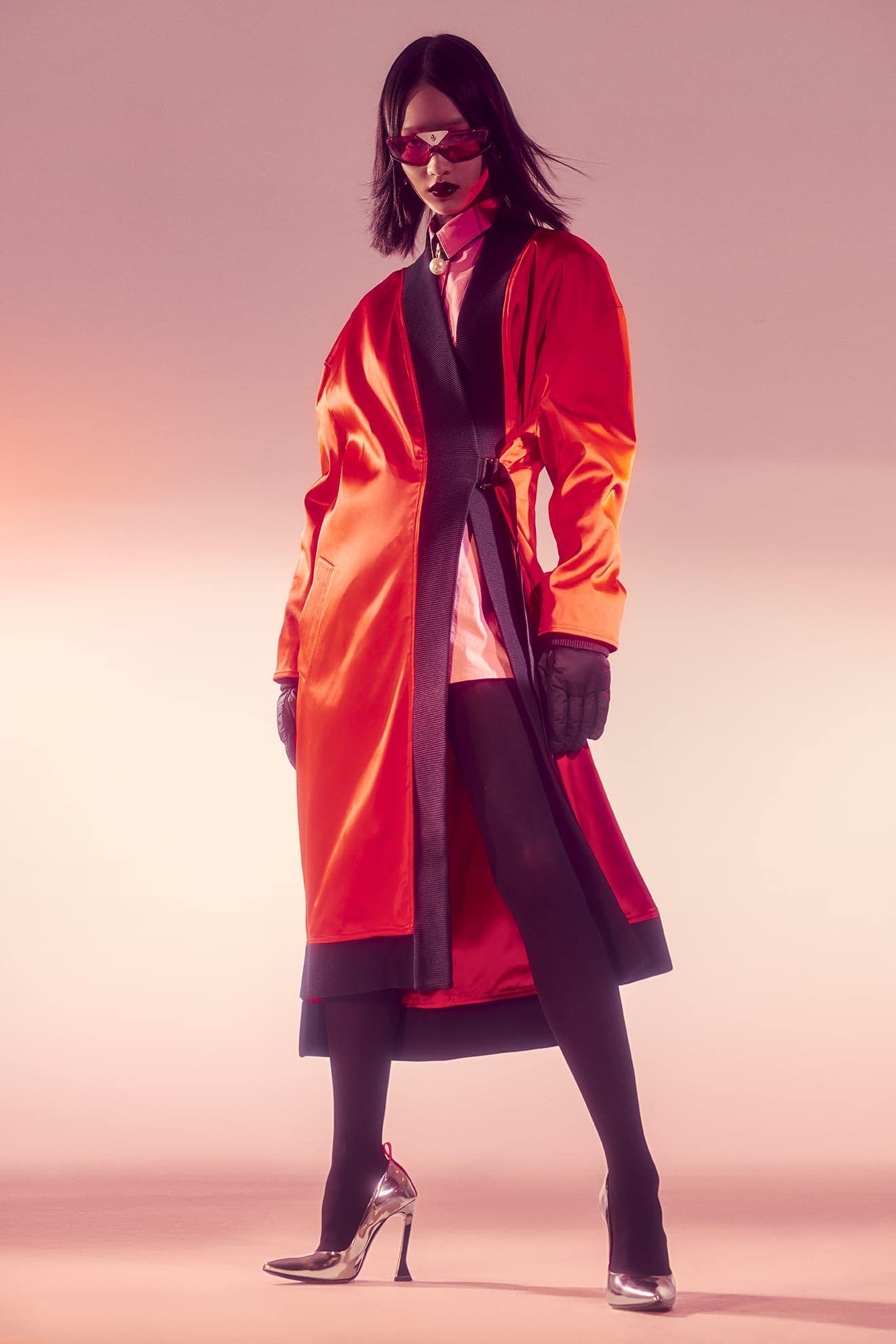 Fashion portrait of an Asian model in an orange satin coat, black tights, and silver heels, posing against a graduated pink backdrop.