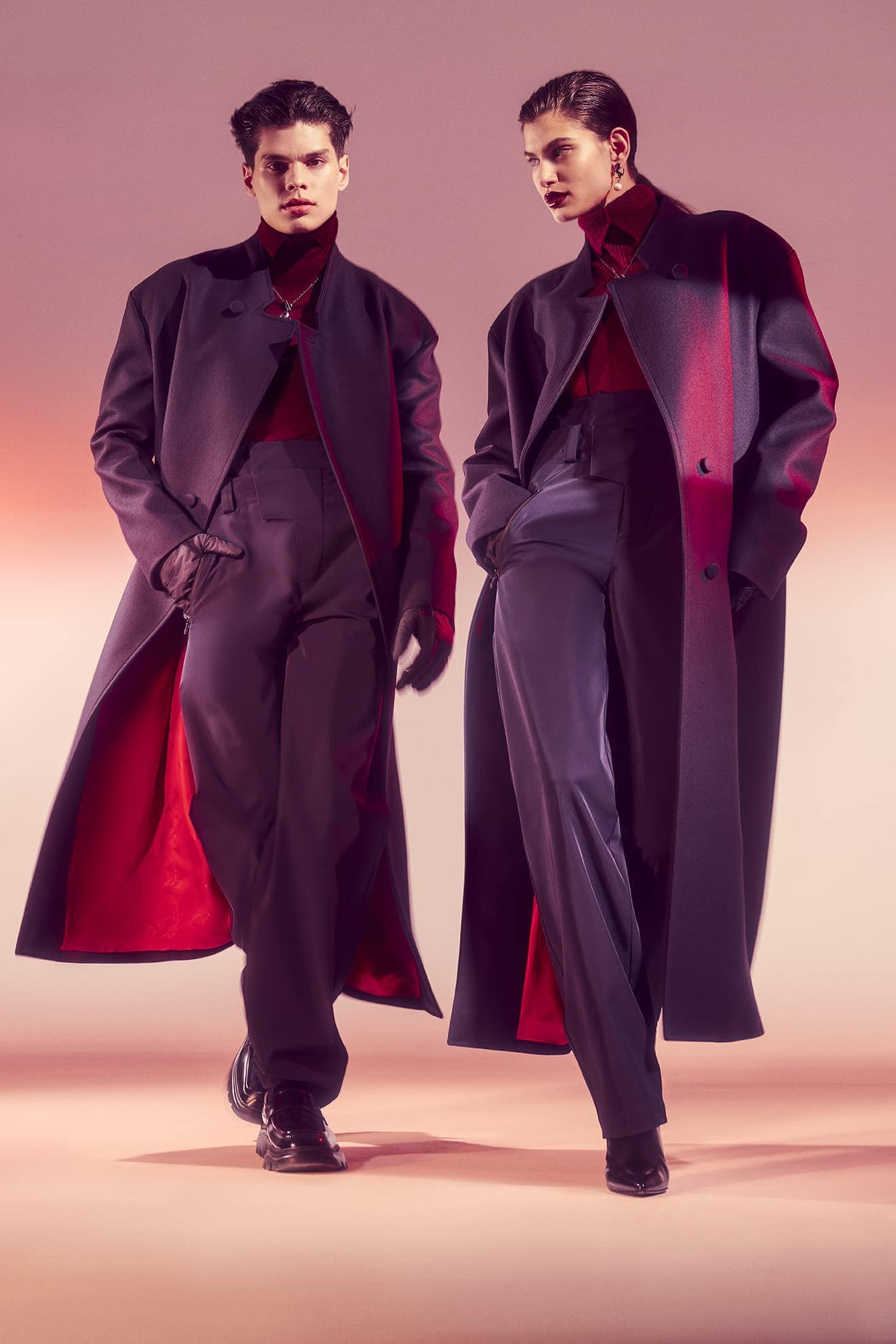 Two fashion models in long coats and dark outfits, posing against a gradient pink background in a studio fashion shot. 