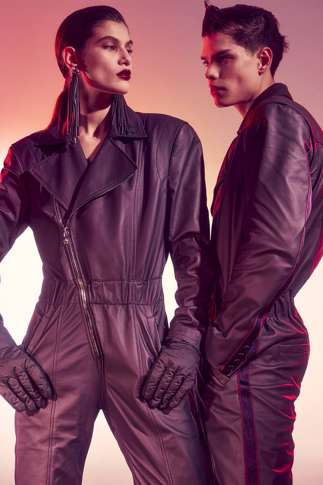 Two fashion models in black leather jumpsuits pose in a studio with gradient red and yellow lighting, showcasing a sleek and edgy aesthetic.