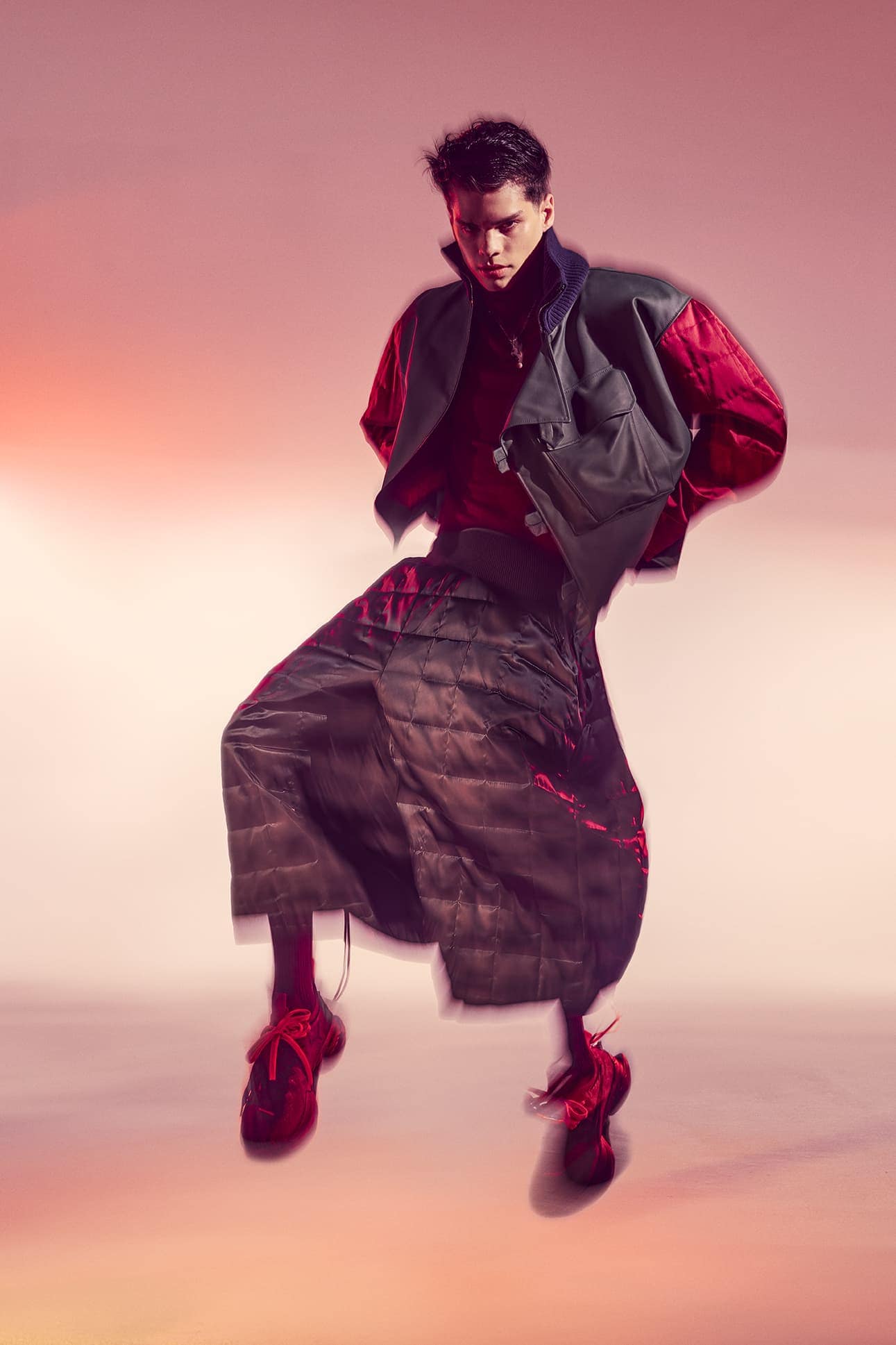 Fashion shot of a young adult man in mid-air wearing a red shirt, vest, quilted pants, and red sneakers against a gradient background.