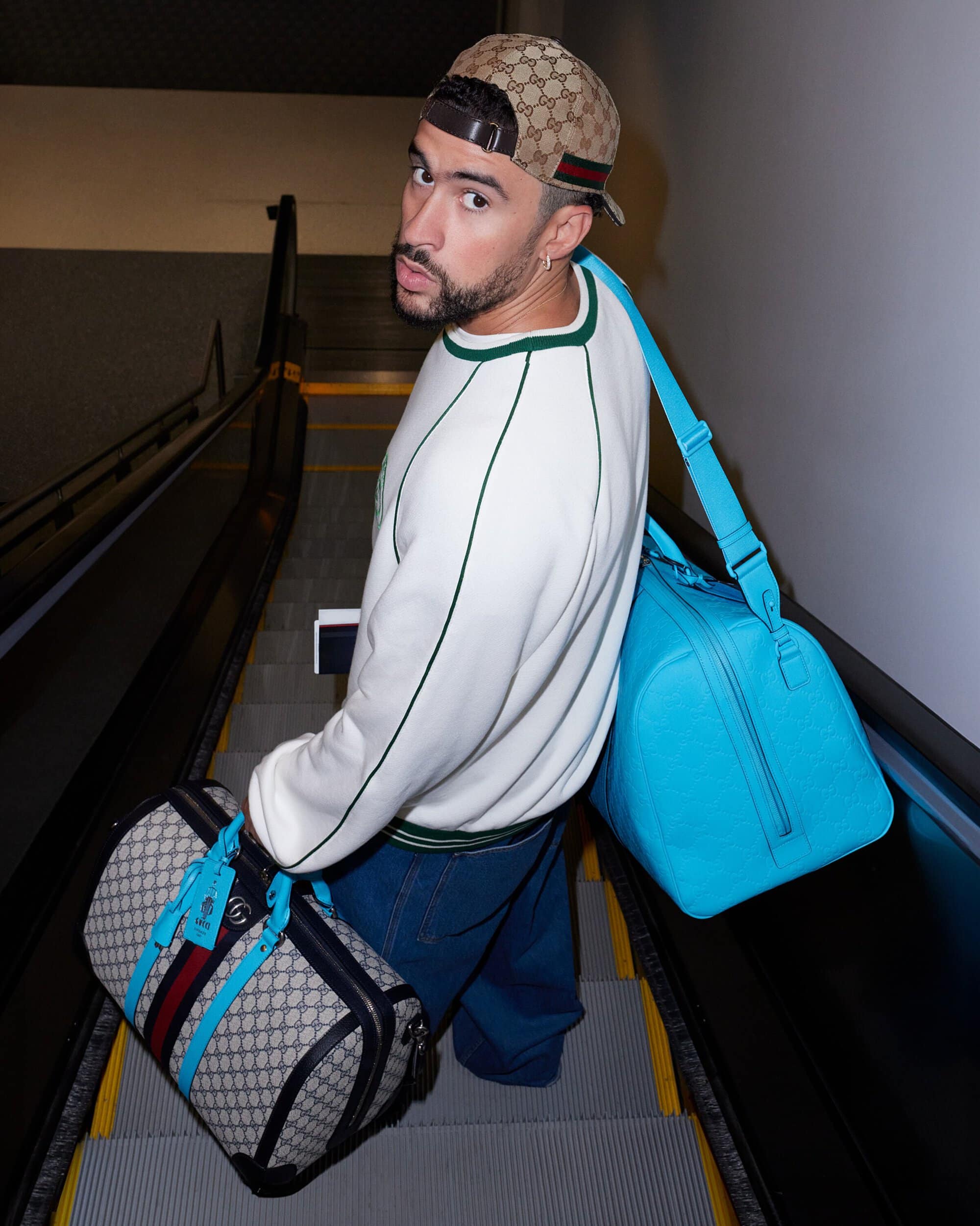 Bad Bunny wearing a Gucci hat and carrying two bags descends on an escalator in an indoor setting with a serious facial expression.
