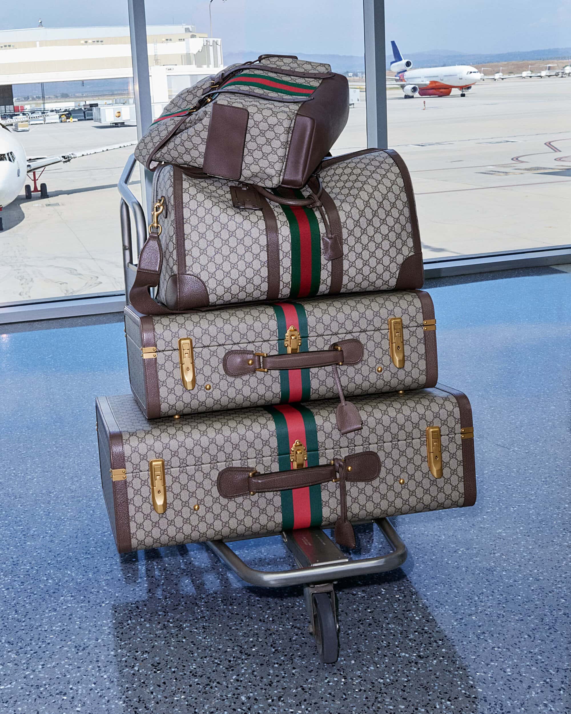 Gucci luggage stacked on a luggage cart in an airport terminal, visible airplanes, bags featuring the Gucci monogram pattern.