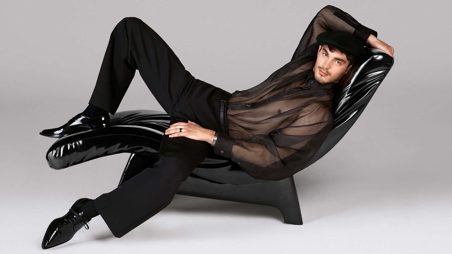 Adult male reclining in a high-gloss black chair. The male is wearing a semi-sheer shirt, trousers, cap, and shiny dress shoes, in studio lighting.